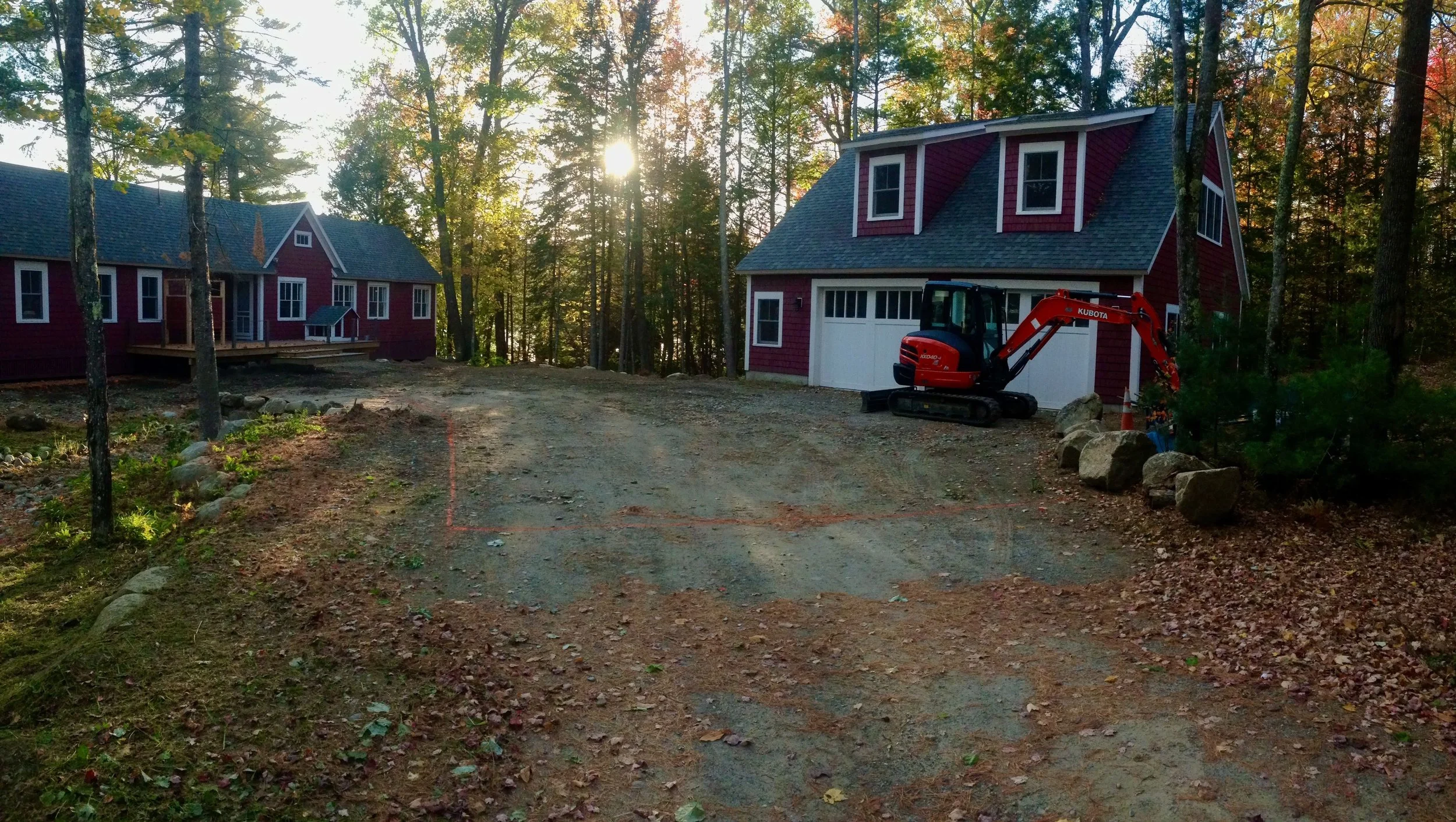 Two red houses under construction surrounded by trees, sun setting in the background, with a dirt clearing and construction equipment in the foreground.