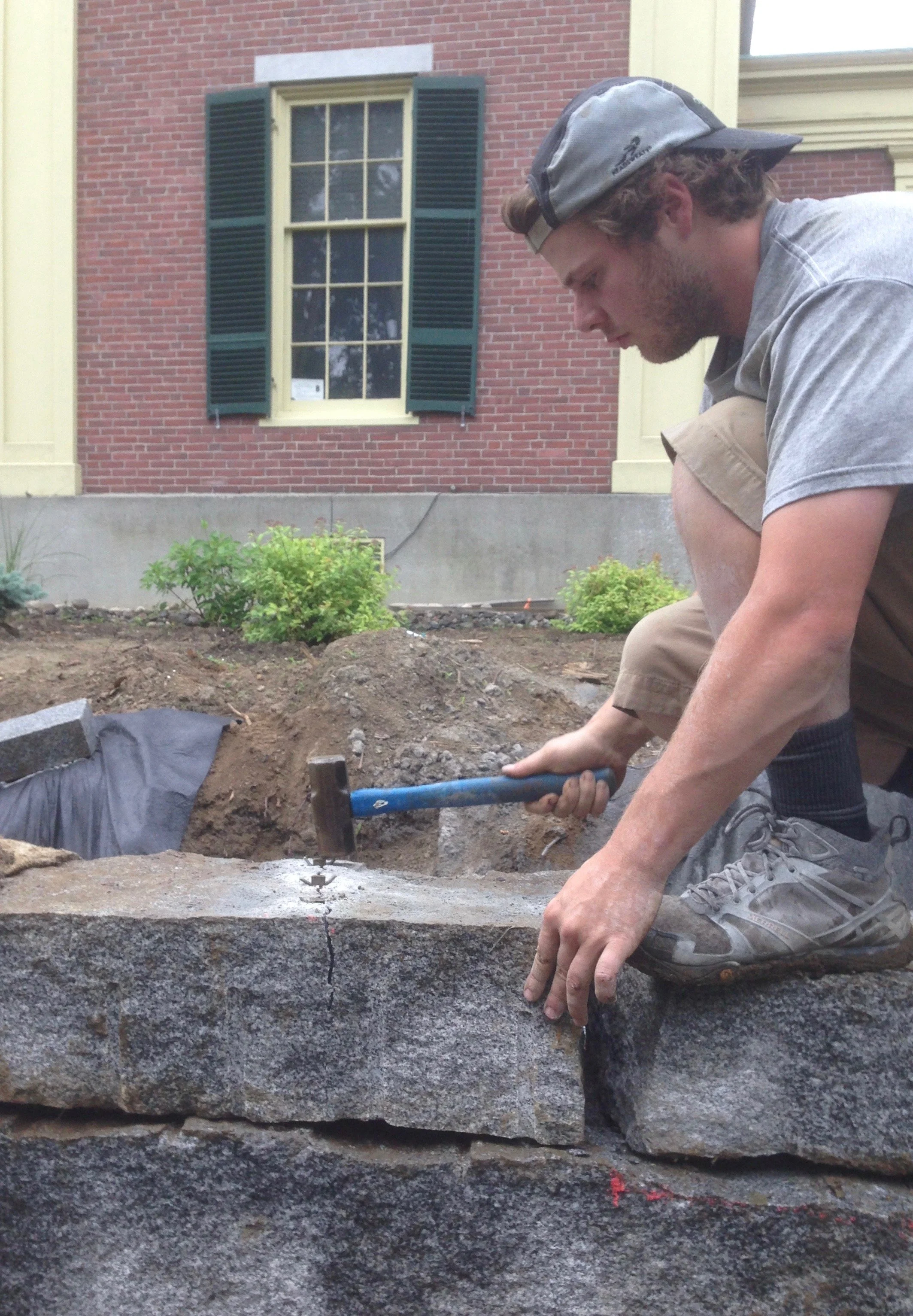 Man kneeling and working on a stone monument or sidewalk, holding a hammer, with a red brick house and window with green shutters in the background.
