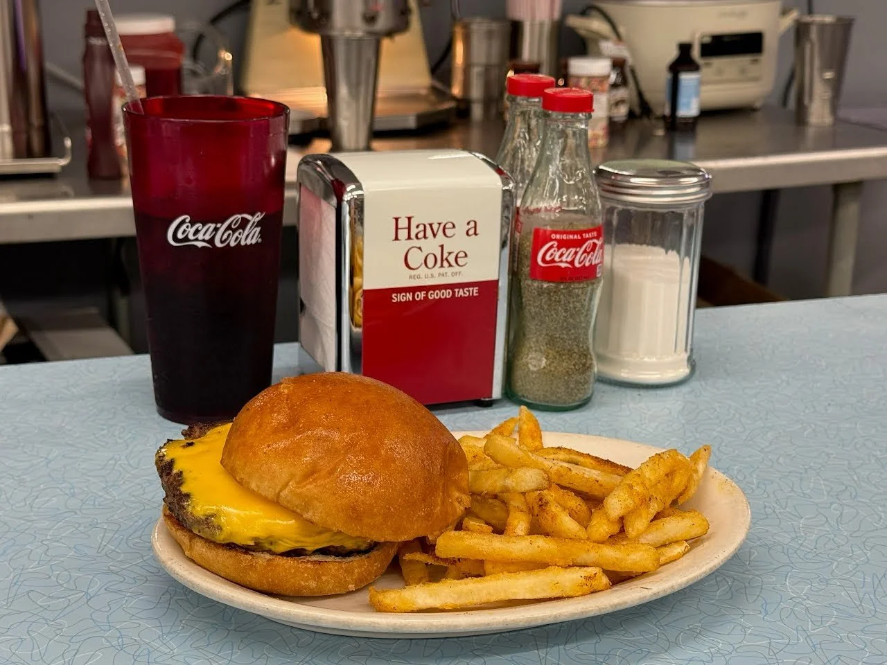 A cheeseburger with fries served on a white plate, a Coca-Cola drink in a tall red glass, and condiments including ketchup, salt, and pepper on a light blue table.