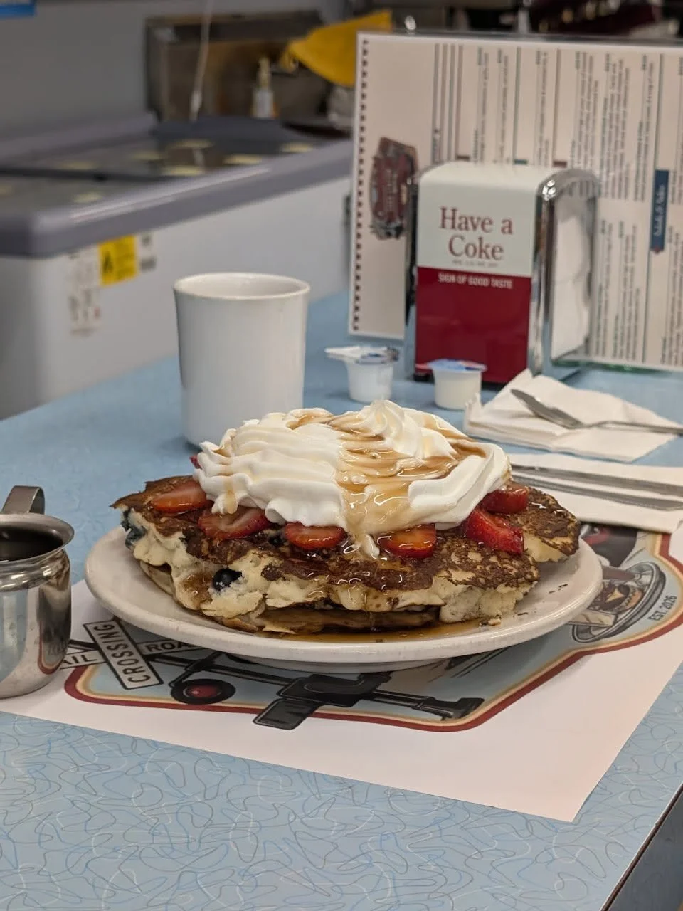 A plate of breakfast food featuring pancakes topped with whipped cream, sliced strawberries, and syrup on a diner table. In the background are a white mug, small creamer cups, a utensil wrapped in a napkin, and a safety pin. A menu and a vintage Coca-Cola box with the words "Have a Coke" are also visible.