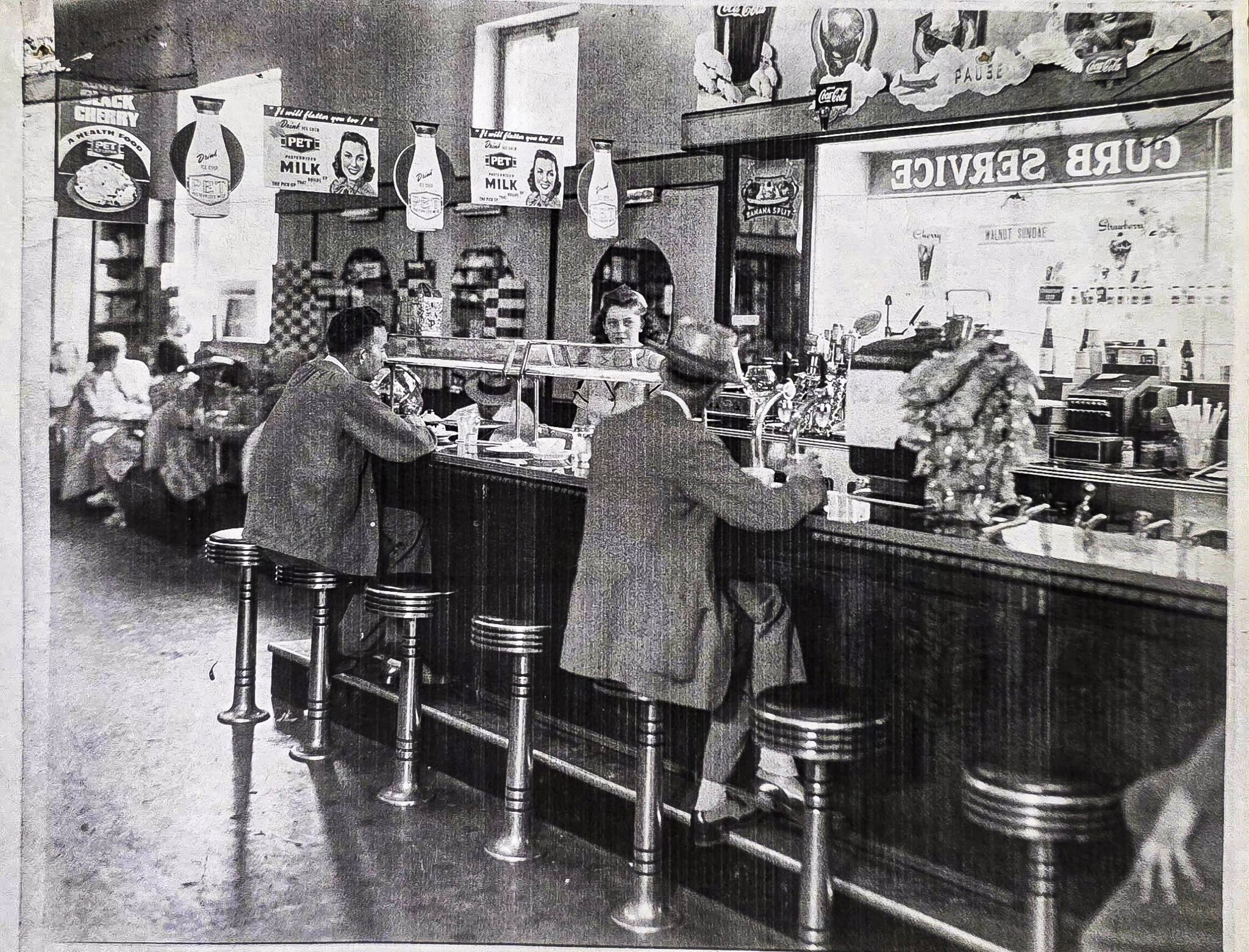 A vintage black and white photo of a diner or cafe interior, showing a counter with stools, customers, and a woman working behind the counter. There are Coca-Cola signs, advertisements, and menu items on the wall.