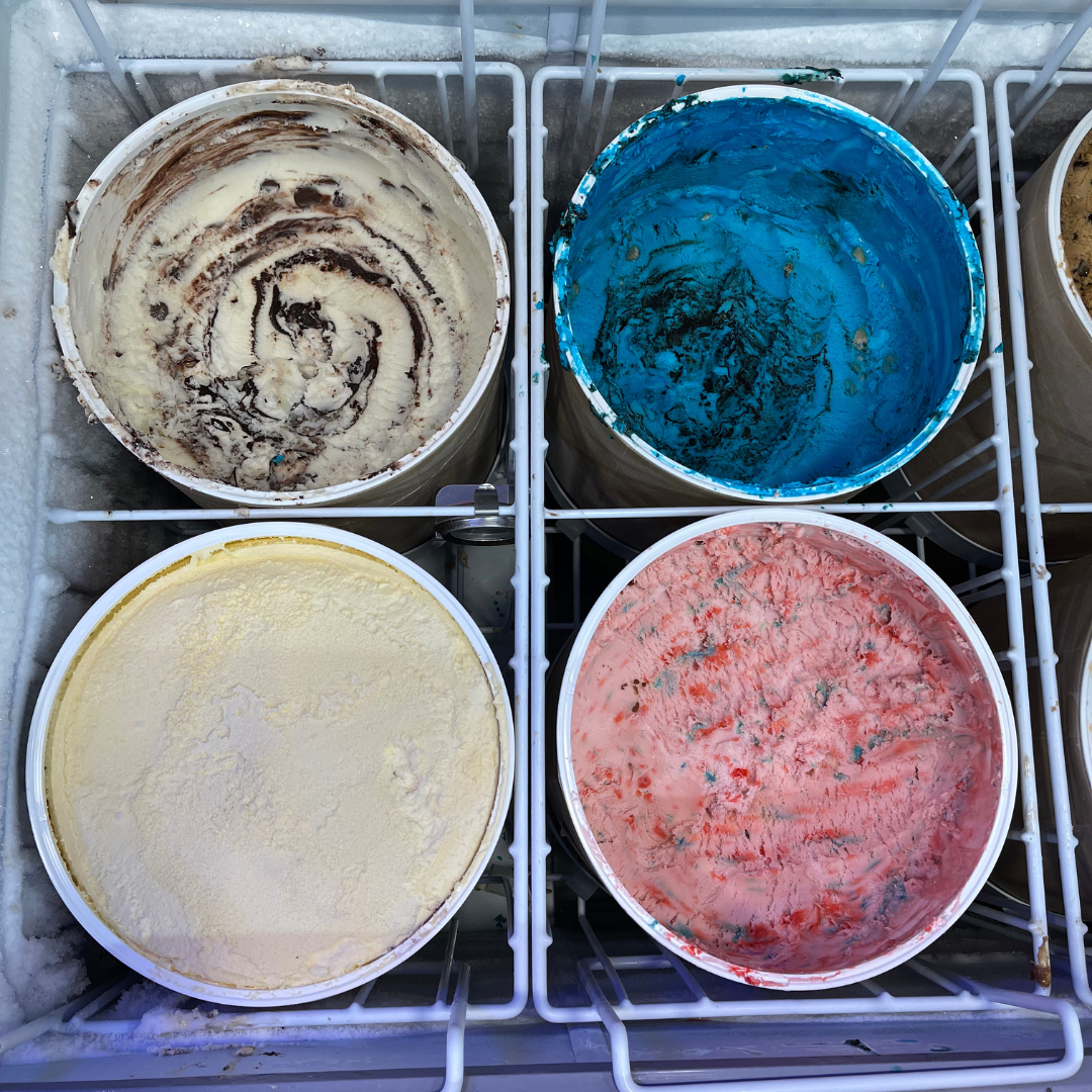 Four open ice cream tubs in a freezer, containing different flavors: chocolate with swirls, blue raspberry, vanilla, and strawberry.
