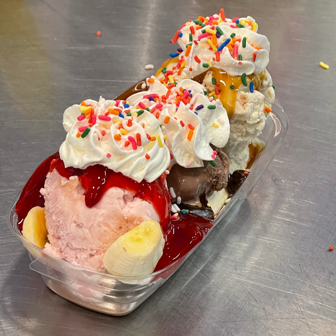 A container of ice cream with different flavors topped with whipped cream and rainbow sprinkles, with banana slices and caramel and berry syrup.