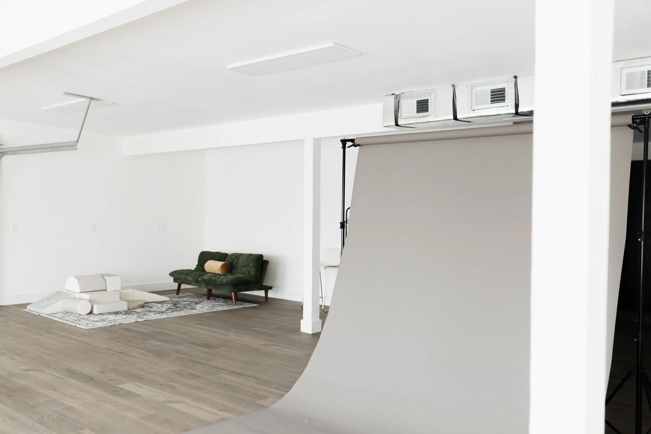 Photography studio with white walls, a green sofa, a patterned rug, a backdrops, and studio lighting equipment.