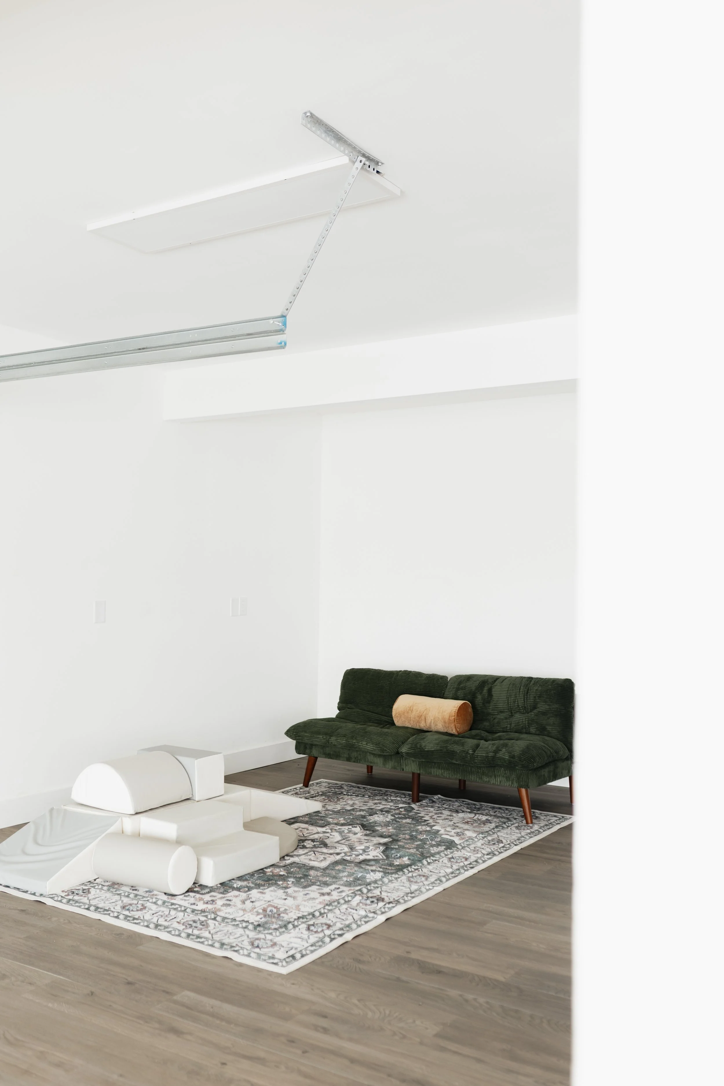 A modern living room with white walls and a dark wood floor, featuring a green velvet sofa with a beige pillow, a light-colored rug, and an abstract white sculpture on the floor. The ceiling has an open metal framework for a ceiling lift or track system.