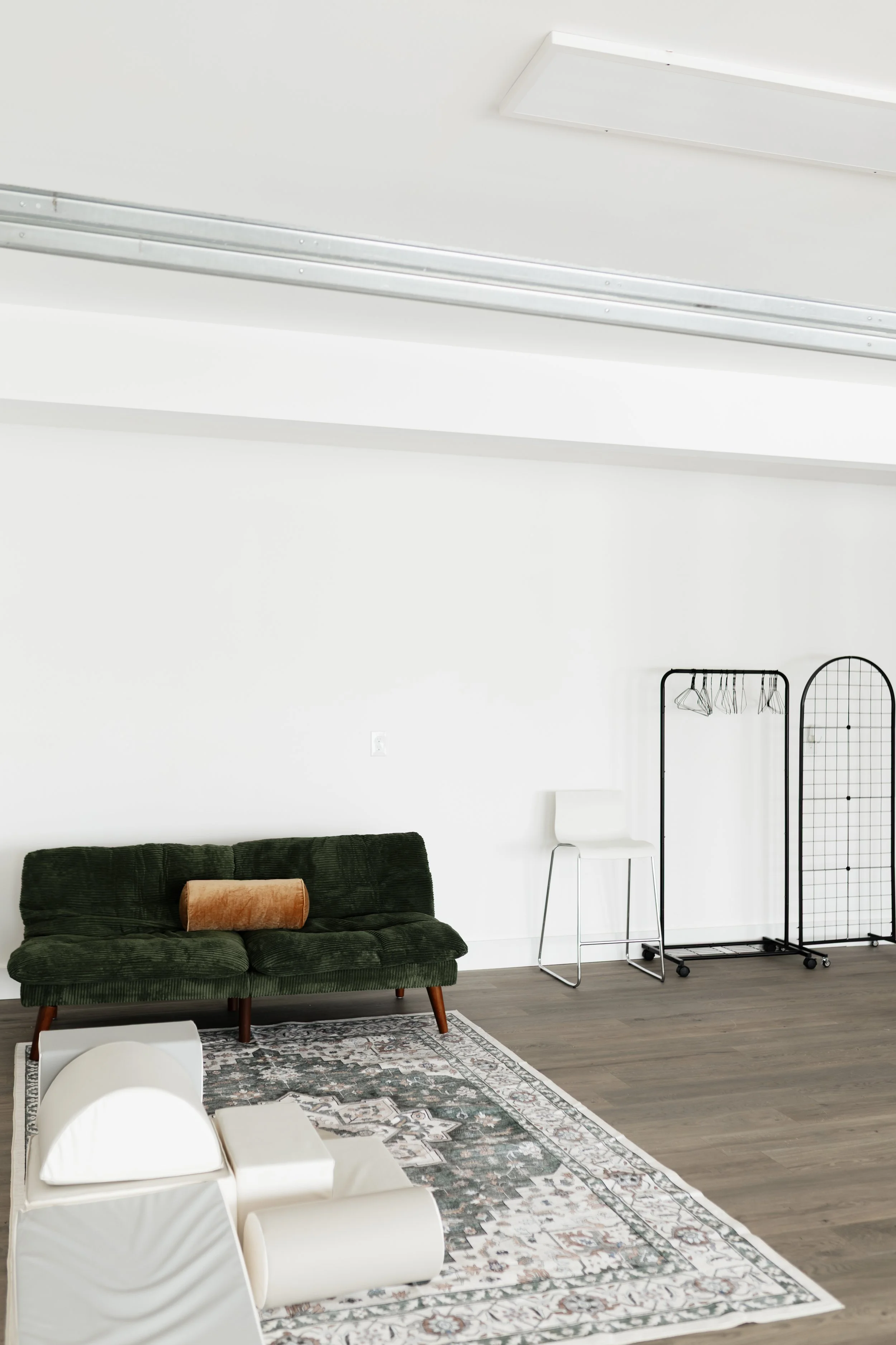 Minimalist living room with white walls, a green velvet sofa with wooden legs and a pillow, a white area rug with floral pattern, a black metal clothing rack with hangers, a white chair, a small white side table, and a black wire screen divider.