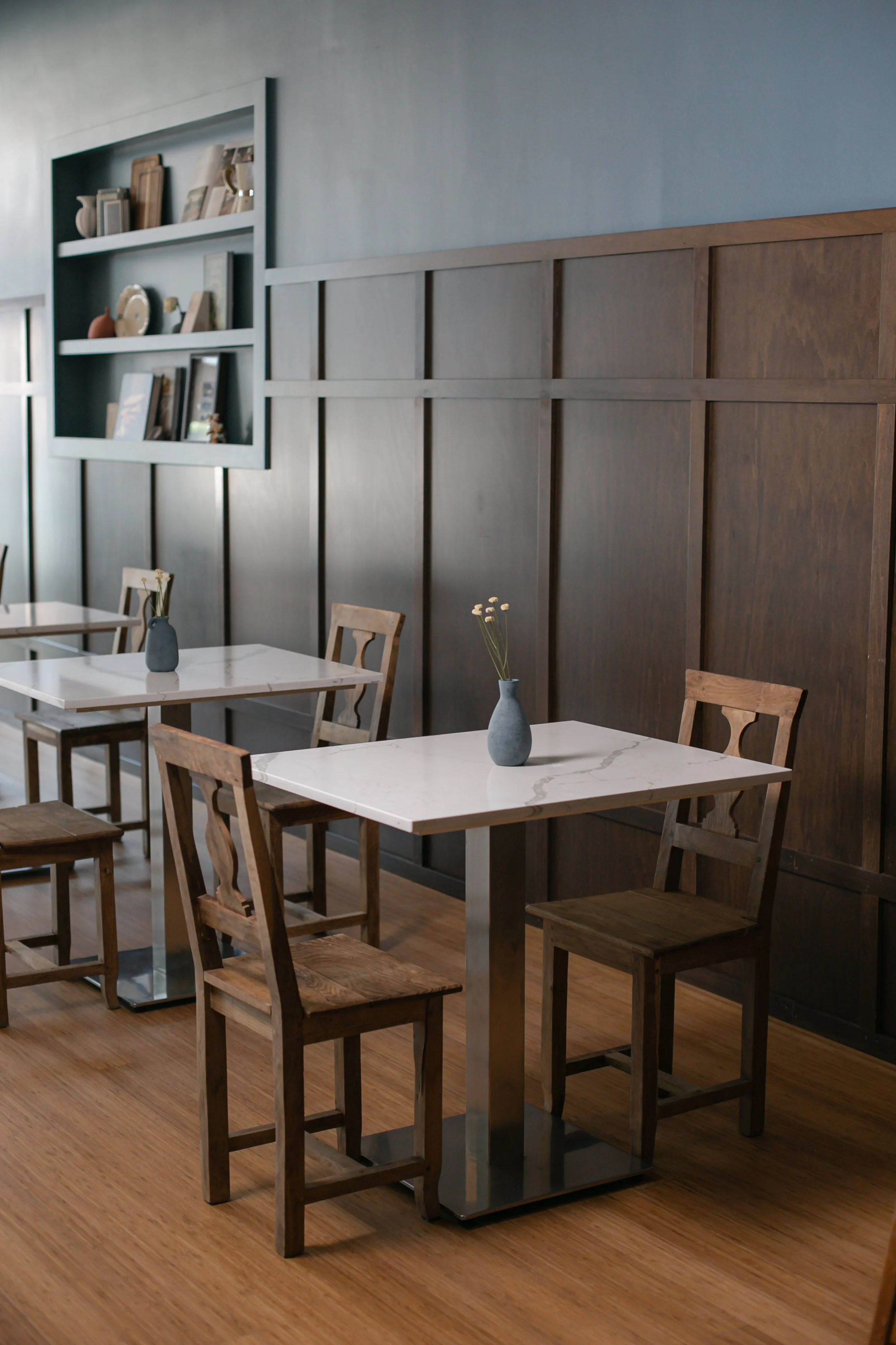 Indoor dining area with multiple white marble tables, wooden chairs, and decorative vases with flowers, with a dark wooden wall and a shelf with decorative objects in the background.