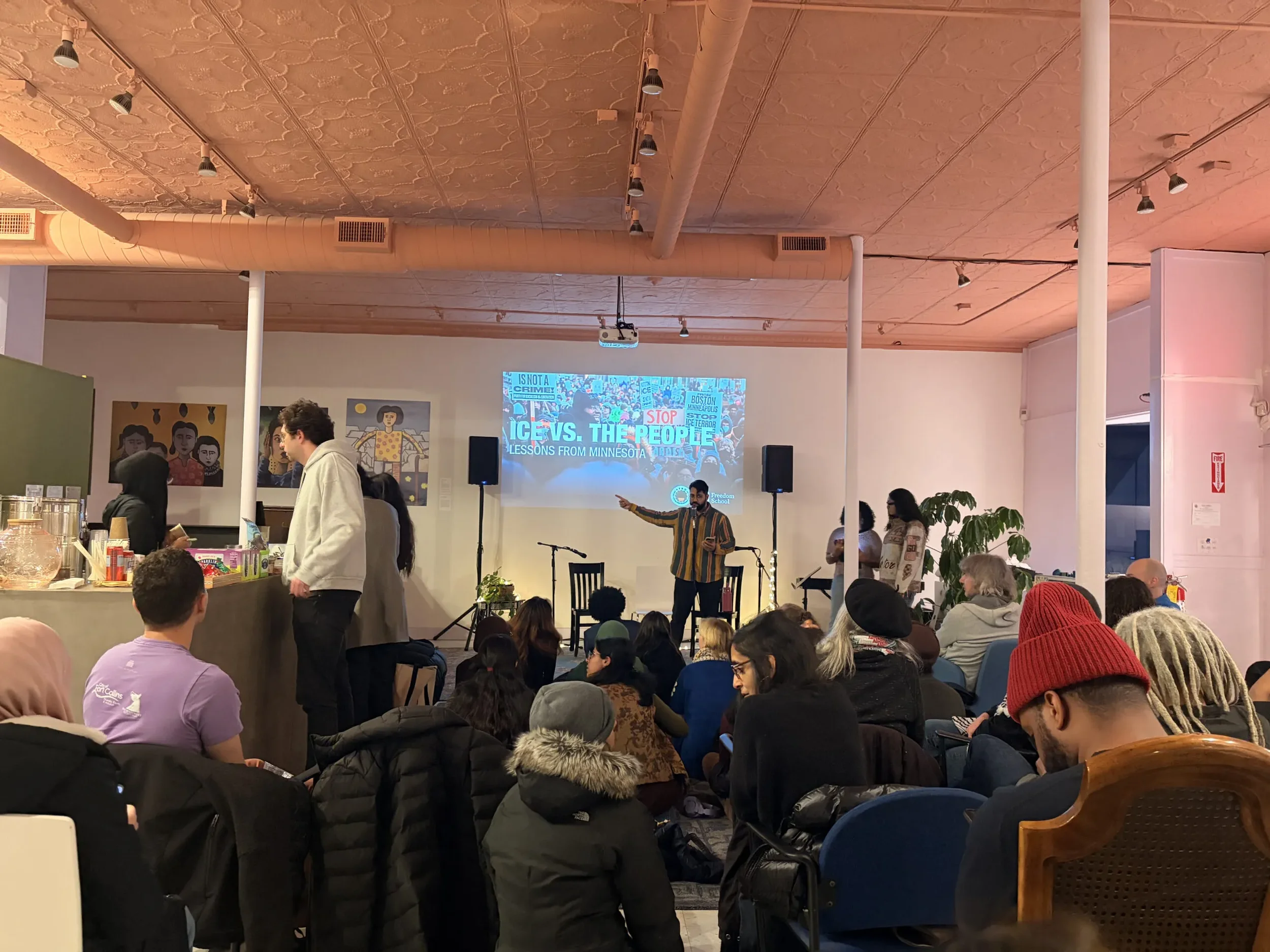A group of people attending a presentation or lecture in an indoor space with a high ceiling and track lighting, seated on chairs and some standing, watching a screen displaying a presentation titled "Ice vs. The People."