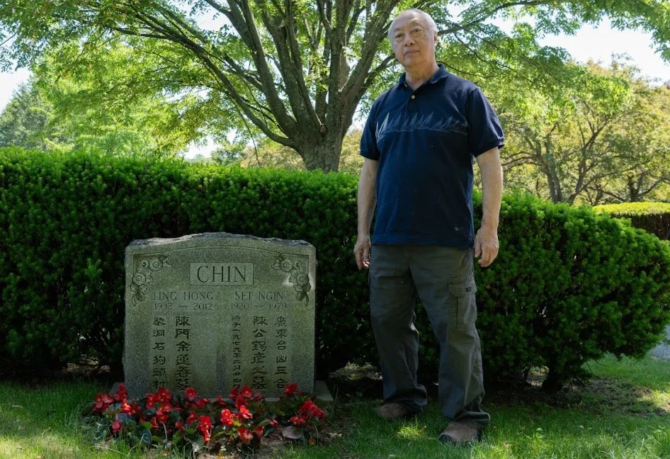 A man standing next to a gravestone with Chinese characters, plants, and trees in a park-like setting.