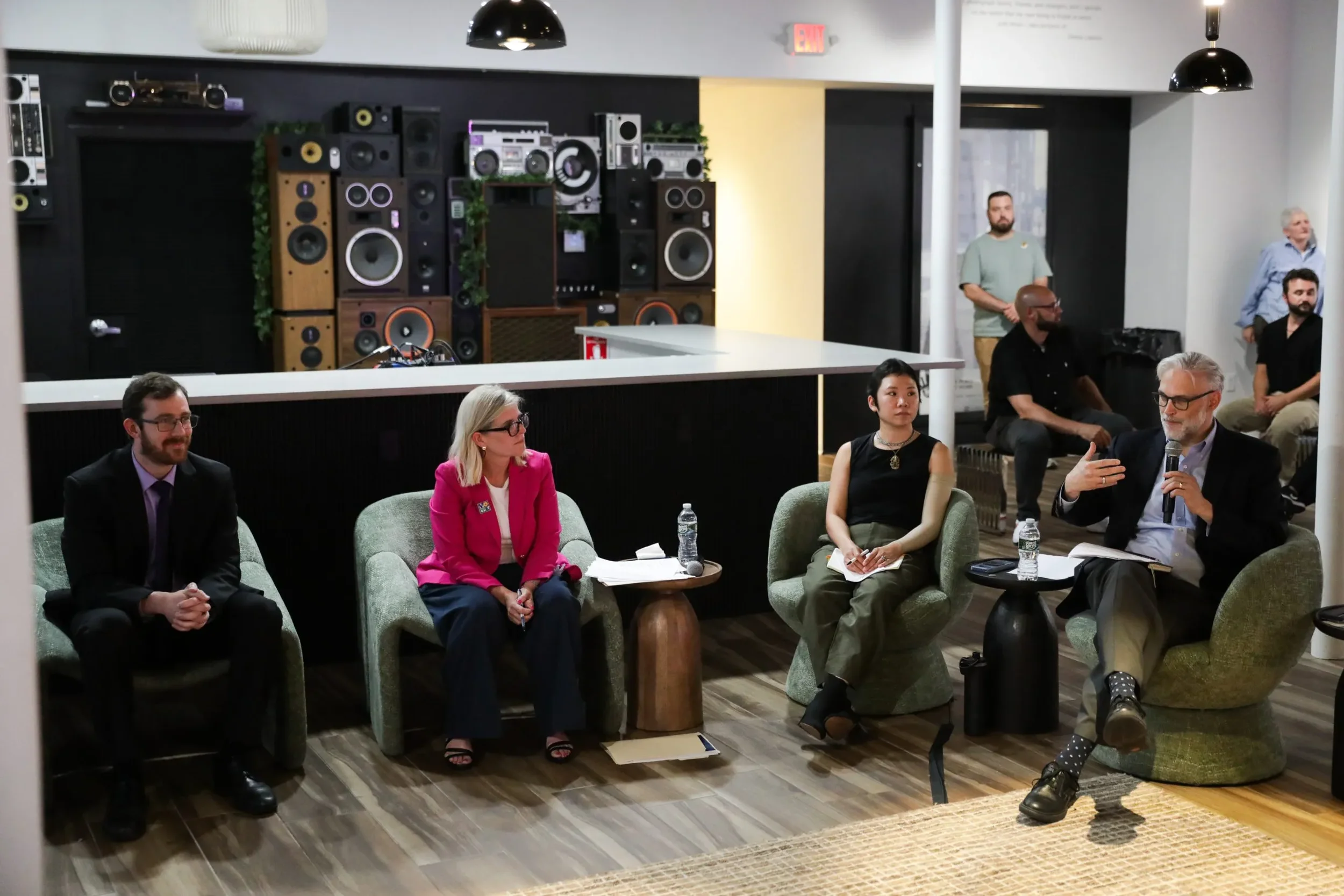 A panel discussion with five people seated on green armchairs, with a man in a suit speaking into a microphone, others with notebooks and water bottles in a room with a modern interior and a large wall of speakers and audio equipment.