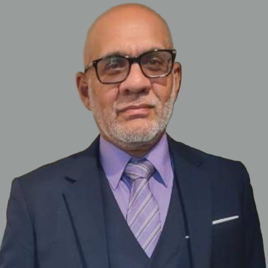 Man with glasses, gray beard, wearing a dark suit, purple shirt, and striped tie, standing against a gray background.