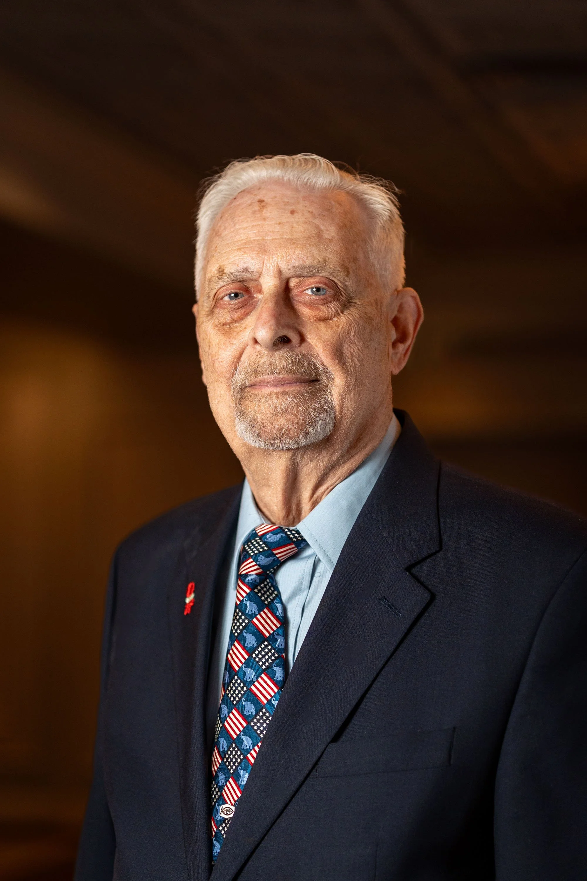 Older man with white hair and beard wearing a dark suit, light blue shirt, and patriotic-themed tie, standing indoors with a wooden ceiling background.