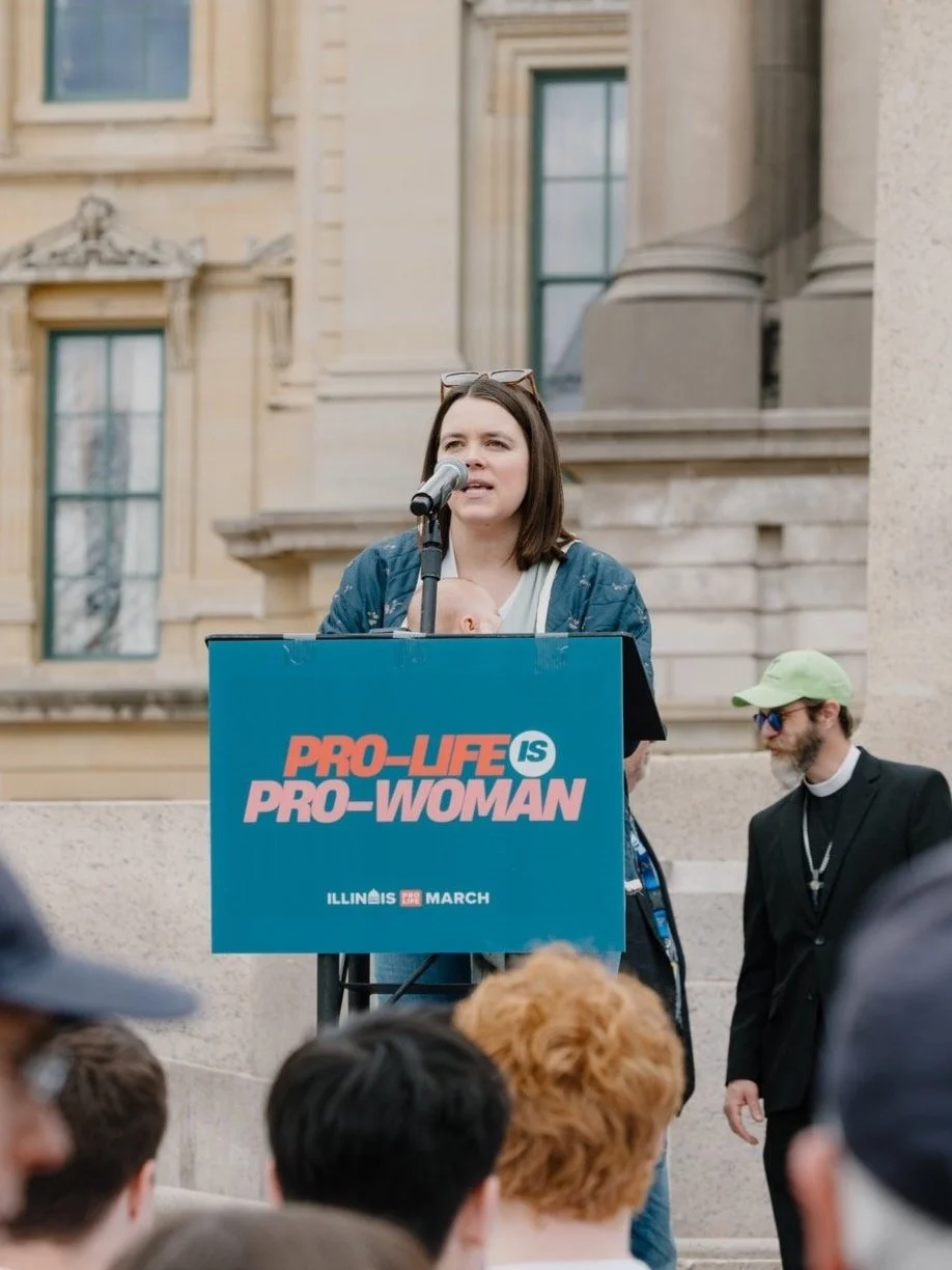 A woman speaking at a pro-life rally on a podium that reads 'Pro-Life is Pro-Woman,' with a crowd in front and a building with columns and windows behind her.