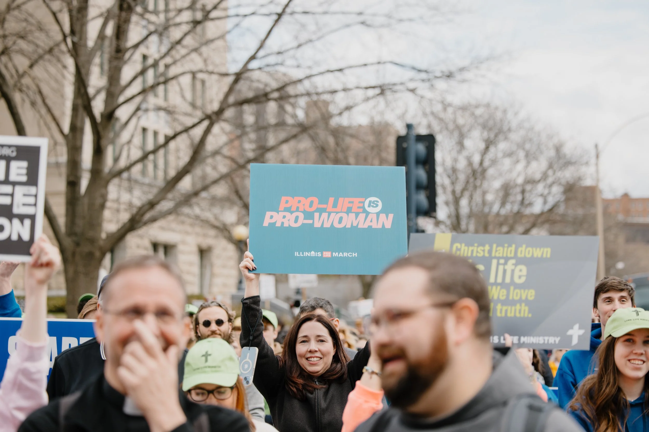 People participating in a protest or march outdoors, holding signs with messages supportive of pro-life and pro-woman viewpoints, with trees and buildings in the background.