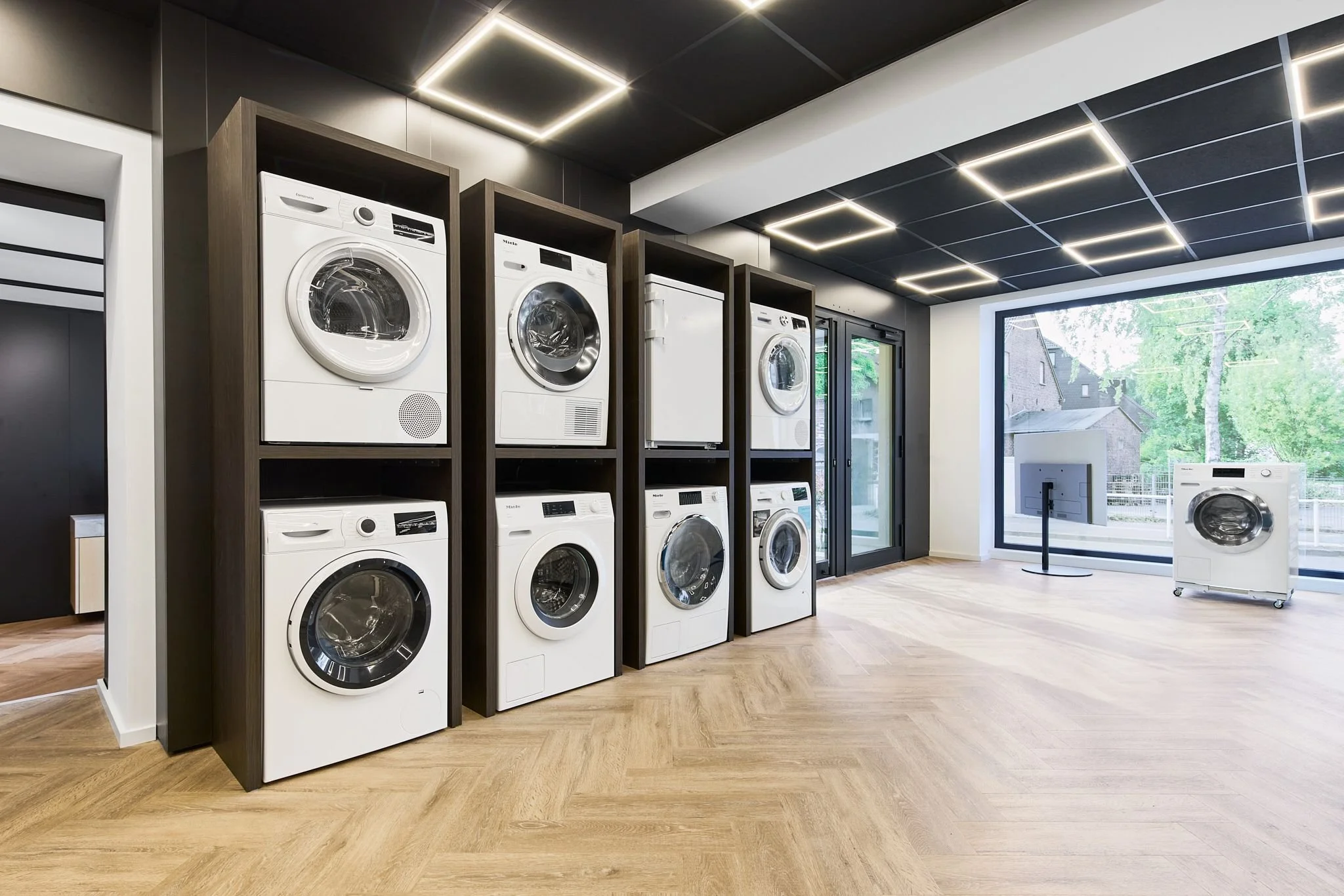 Saubere Waschmaschinen stehen in einem hellen, modernen Ausstellungsraum der Radio Stein in Ratingen. Große Fenster lassen Tageslicht herein.