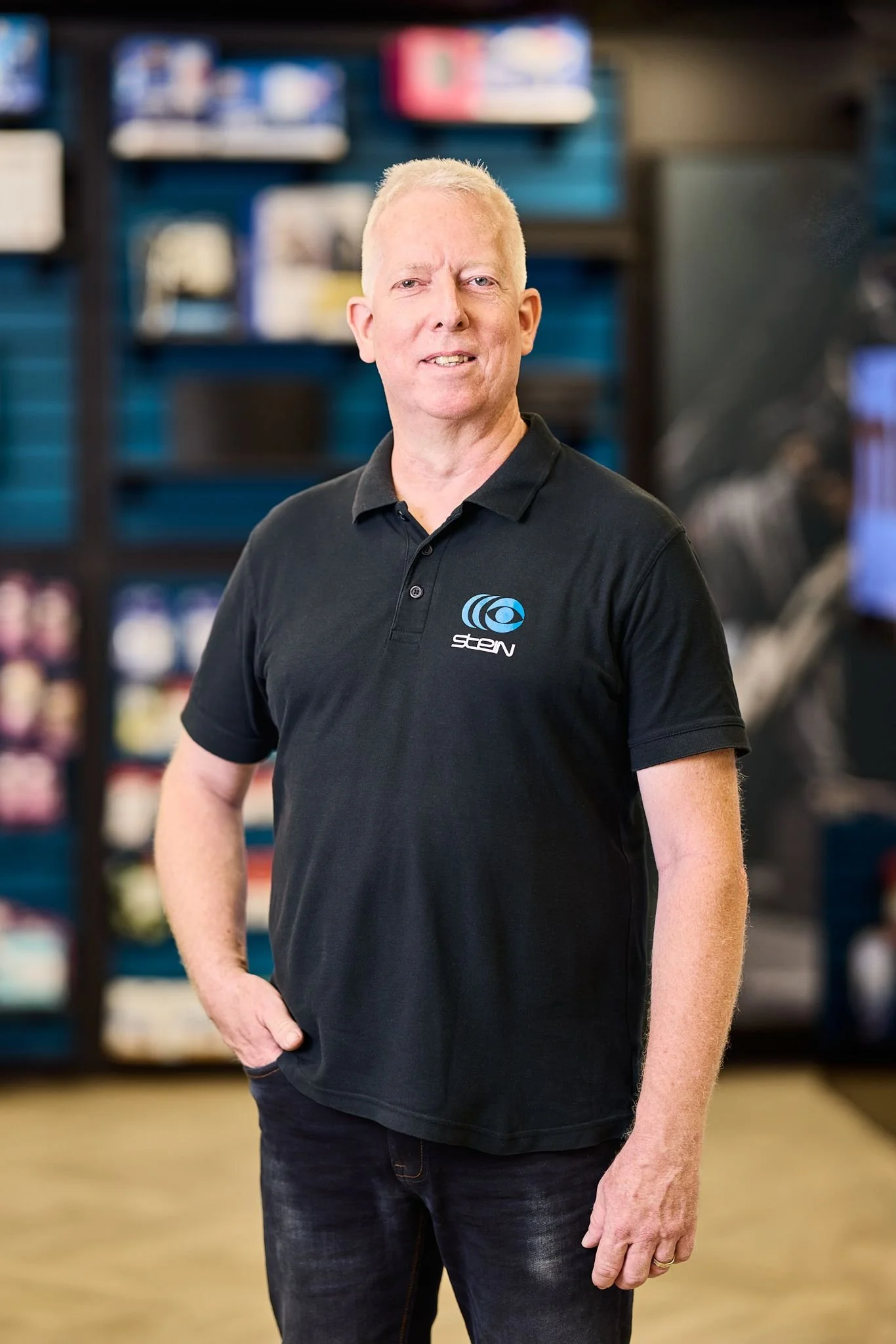 Radio Stein Berater steht in seinem Elektronikfachgeschäft, trägt ein schwarzes Polo-Shirt mit einem Logo auf der linken Brustseite, steht in einem Ladenregal im Hintergrund.