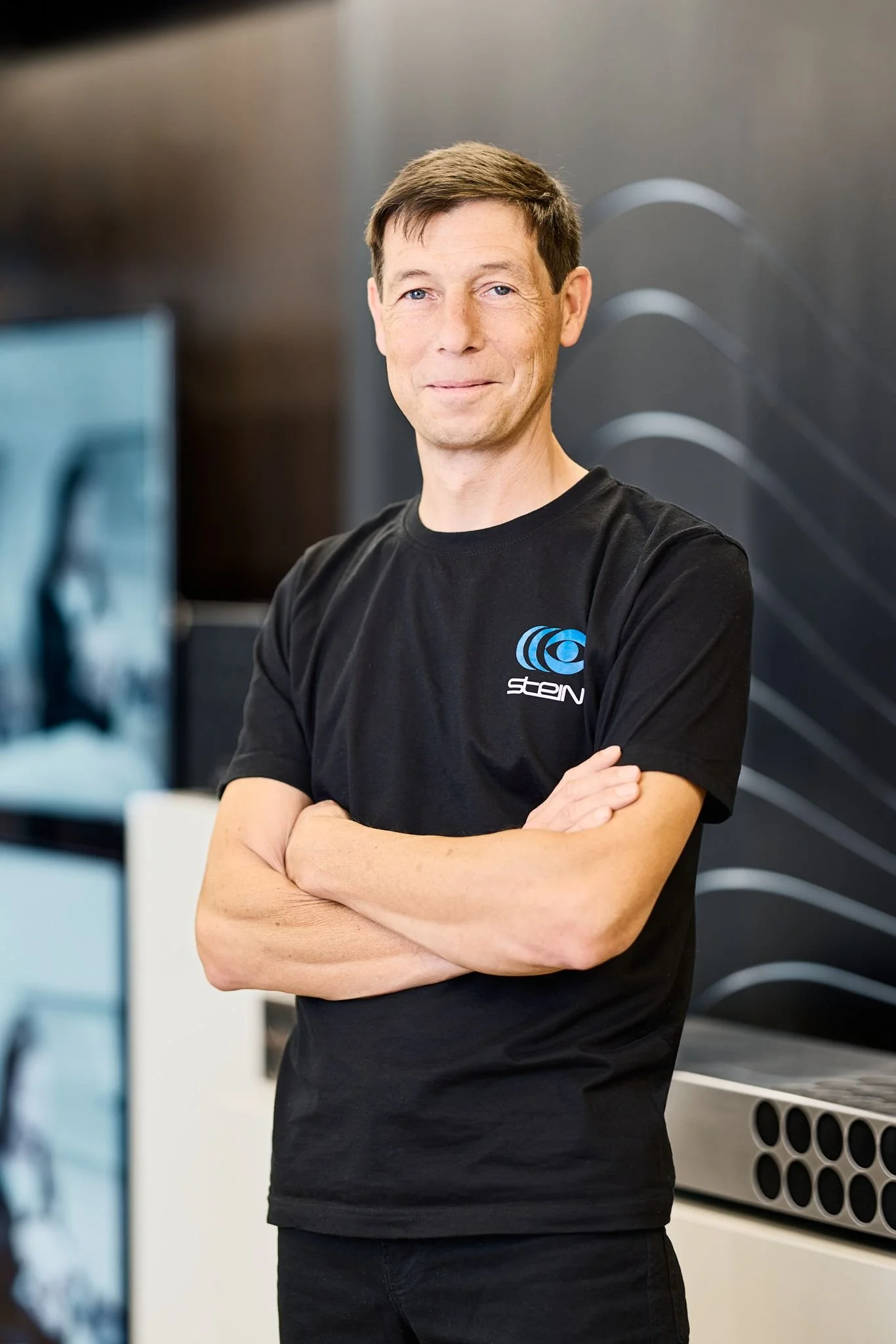 Radio Stein Berater steht in seinem Elektronikfachgeschäft. Er hat kurze, braune Haare, trägt ein schwarzes T-Shirt mit einem blauen Logo auf der Brust und steht mit verschränkten Armen vor einer schwarzen Wand im Büro.