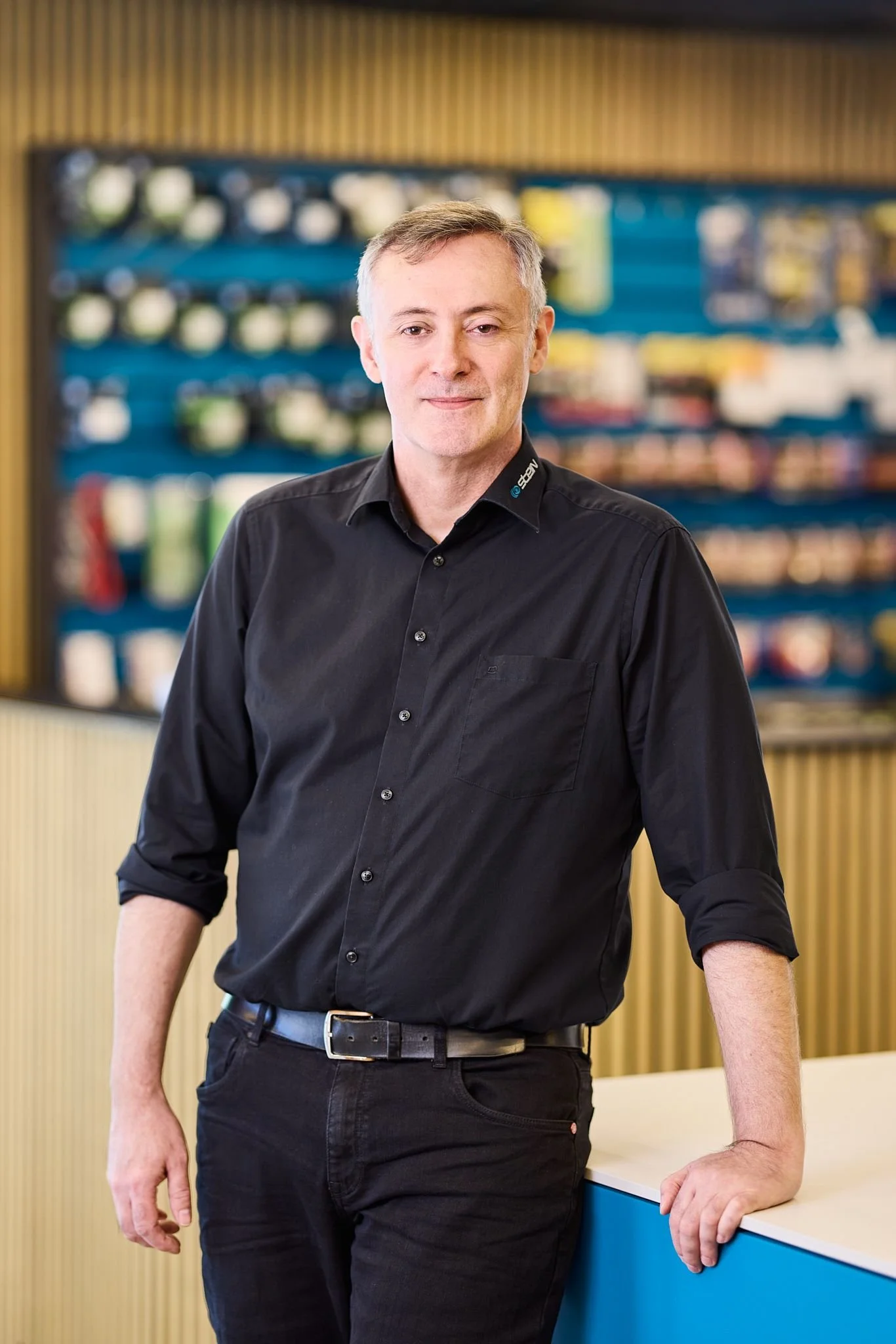Radio Stein Berater steht in seinem Elektronikfachgeschäft vor einer blauen Theke, trägt ein schwarzes Hemd und schwarze Hosen, und lehnt leicht an der Theke.