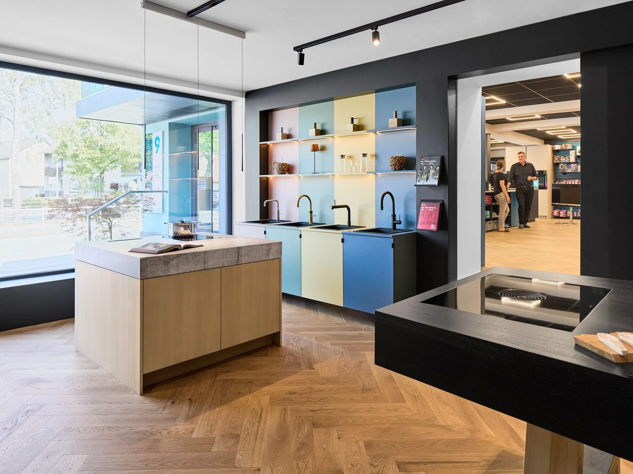 Moderne Kücheneinrichtung im Showroom von Radio Stein in Ratingen mit schwarzem Kochfeld, Waschbecken, farblich gestalteter Wand mit Regalen, mehreren Kochendwasser-Armaturen, und einem großen Fenster mit Blick nach draußen.