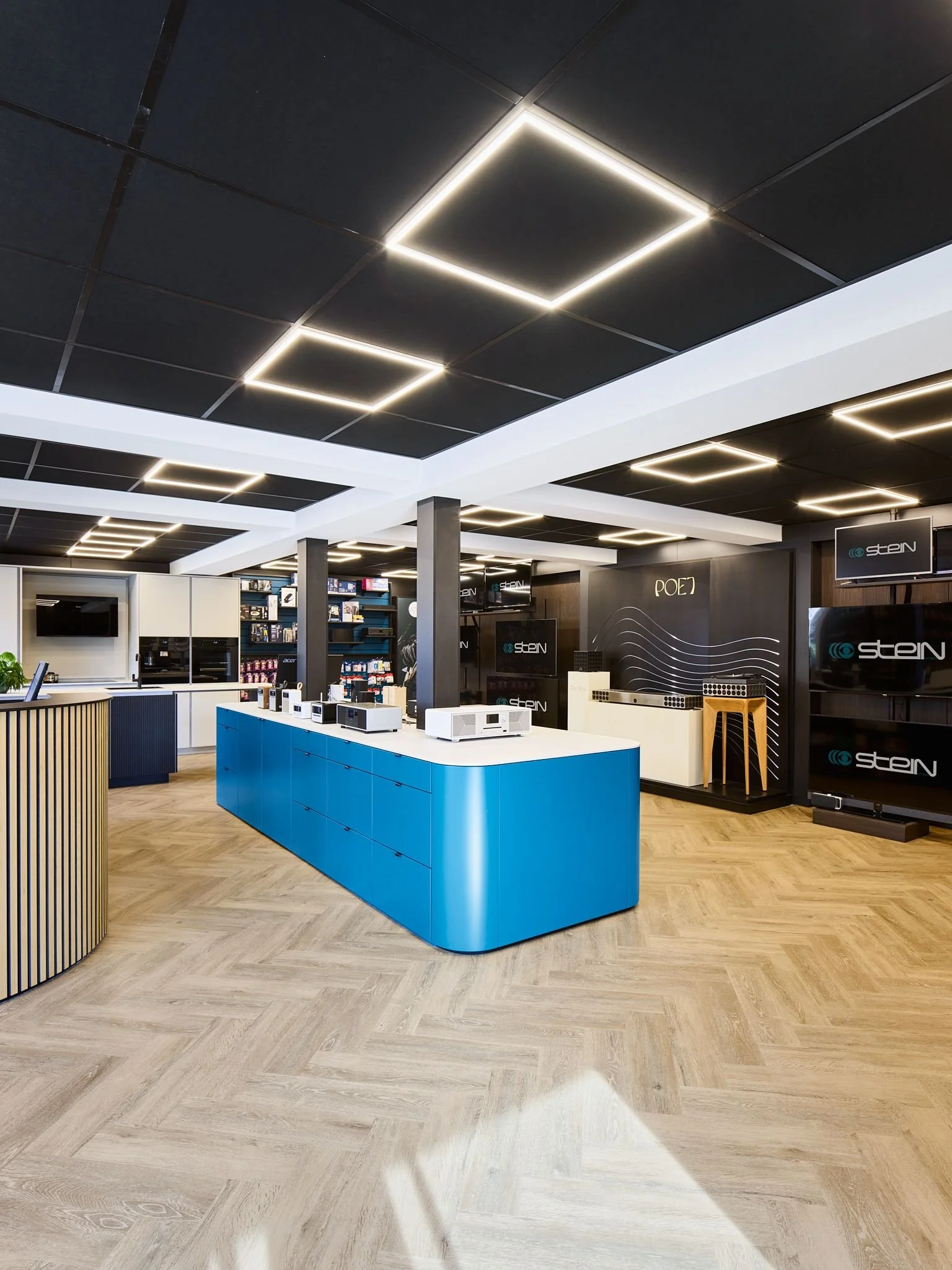 Moderne und freundliche Inneneinrichtung des Technikgeschäfts Radio Stein mit schwarzem Decken mit LED-Lichtern in Form von Quadraten, blauer zentraler Theke, hellen Holzboden, Monitoren und Regalen mit Produkten.