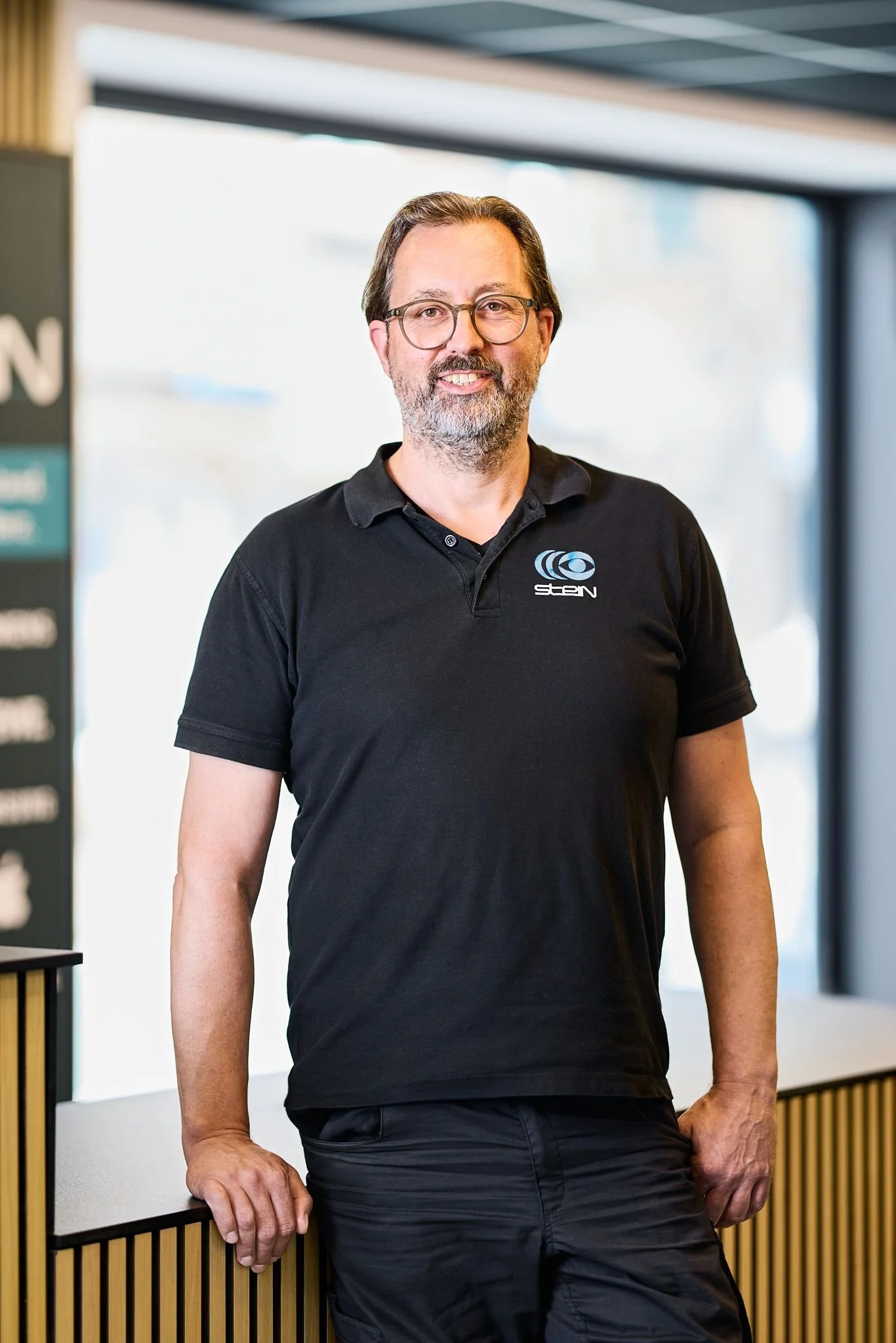 Radio Stein Berater steht in seinem Elektronikfachgeschäft.Er trägt eine Brille in Schwarzshirt steht entspannt an einer Holzbarriere im Innenraum eines modernen Gebäudes.