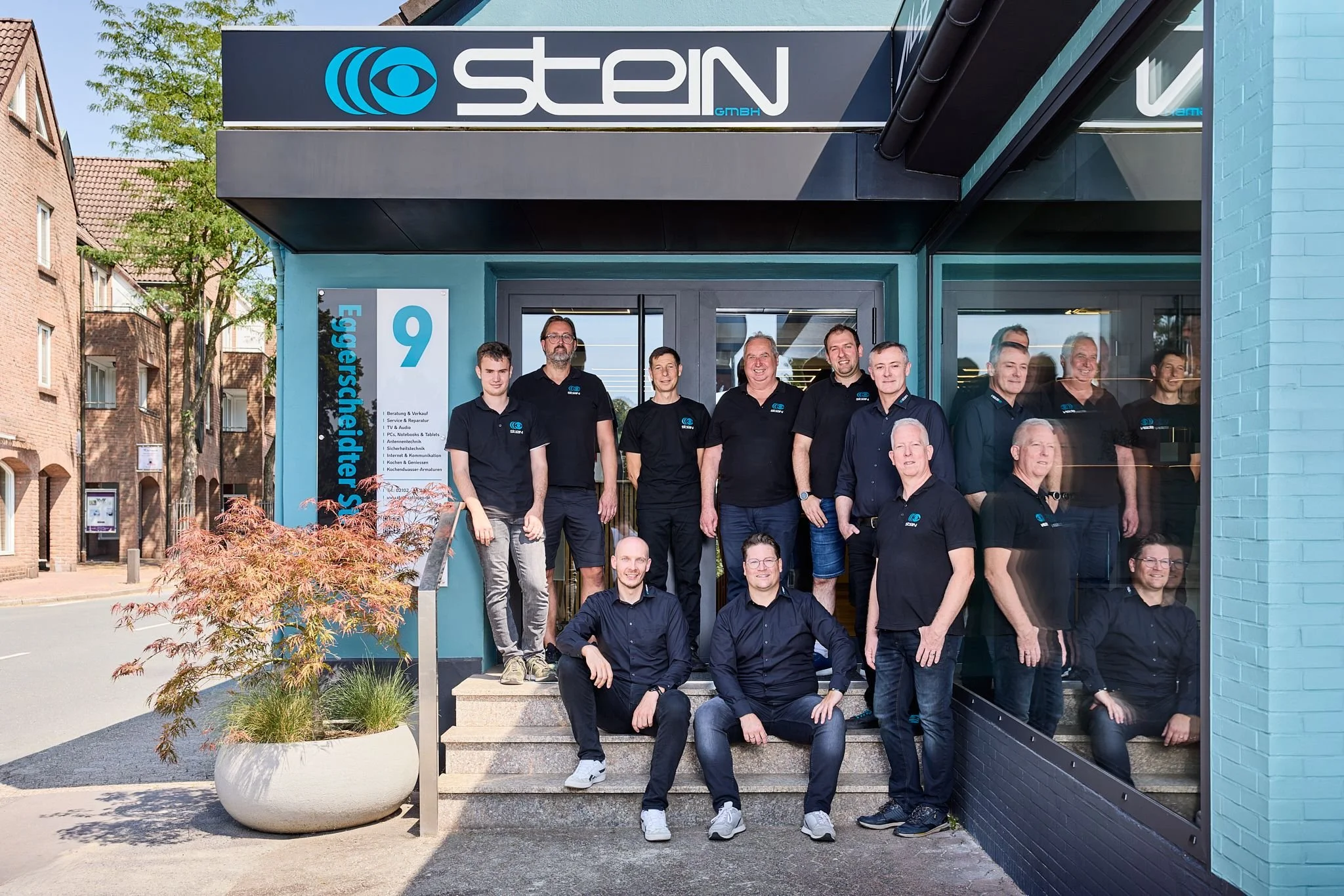 Das Radio Stein Team steht vor dem Ladenlokal in Ratingen. Die Personen tragen schwarze Polohemden mit einem blauen Logo und posieren für das Foto.