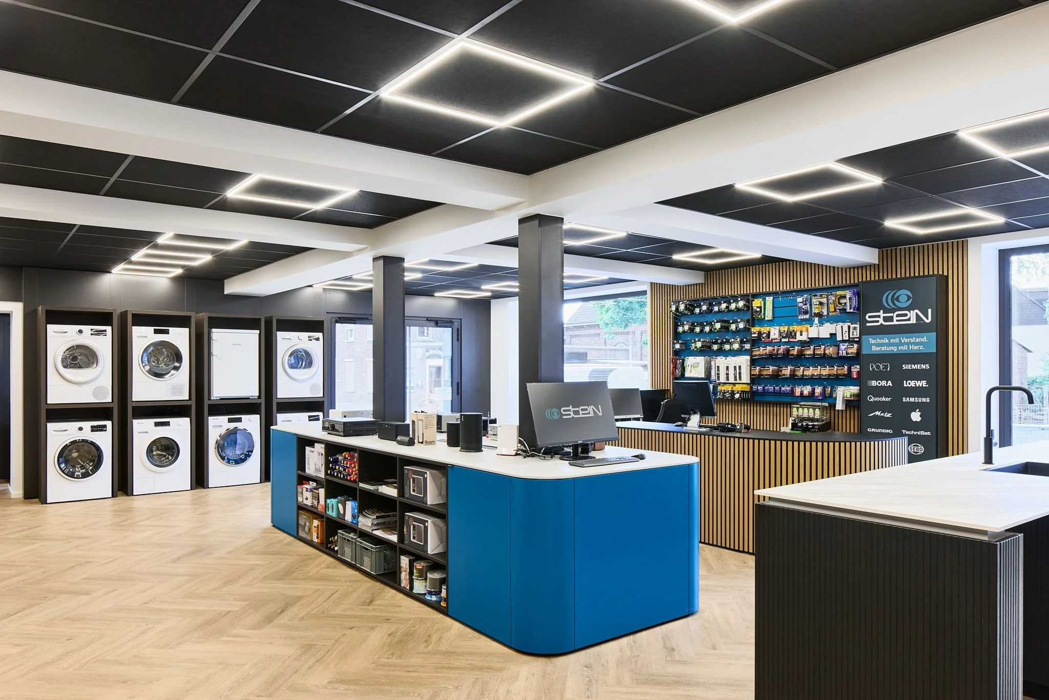 Innenansicht des Elektronik- und Haushaltswarengeschäfts Radio Stein in Ratingen mit Waschmaschinen, einem Verkaufsraum mit Beratungstresen, vielen Produkten und Beleuchtung in modernem Design.