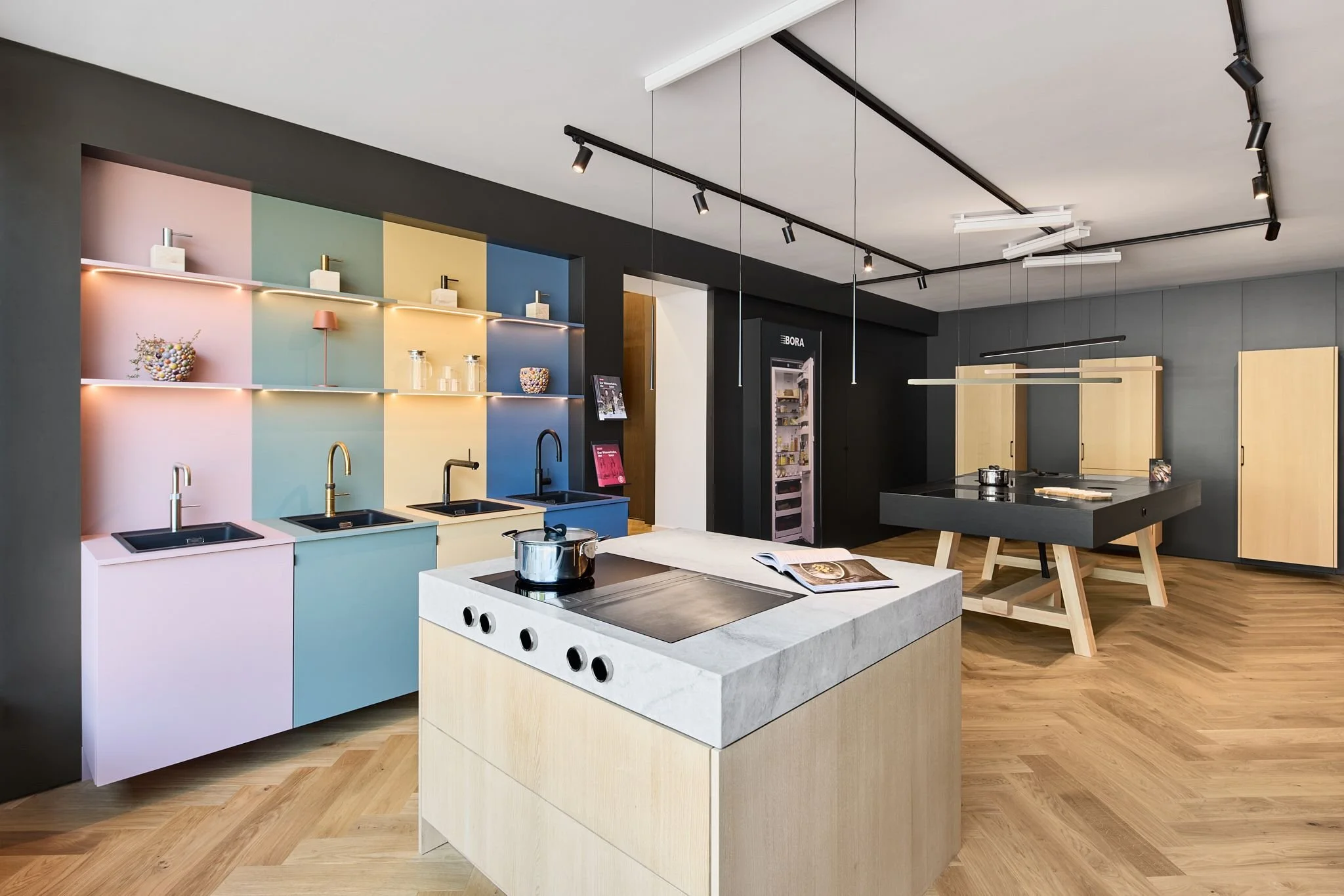 Moderner Küchen-Showroom der Radio Stein GmbH in Ratingen mit farbigen Schränken in Pastellfarben, einer Kochinsel aus Holz und einem schwarzen Esstisch mit Stühlen.