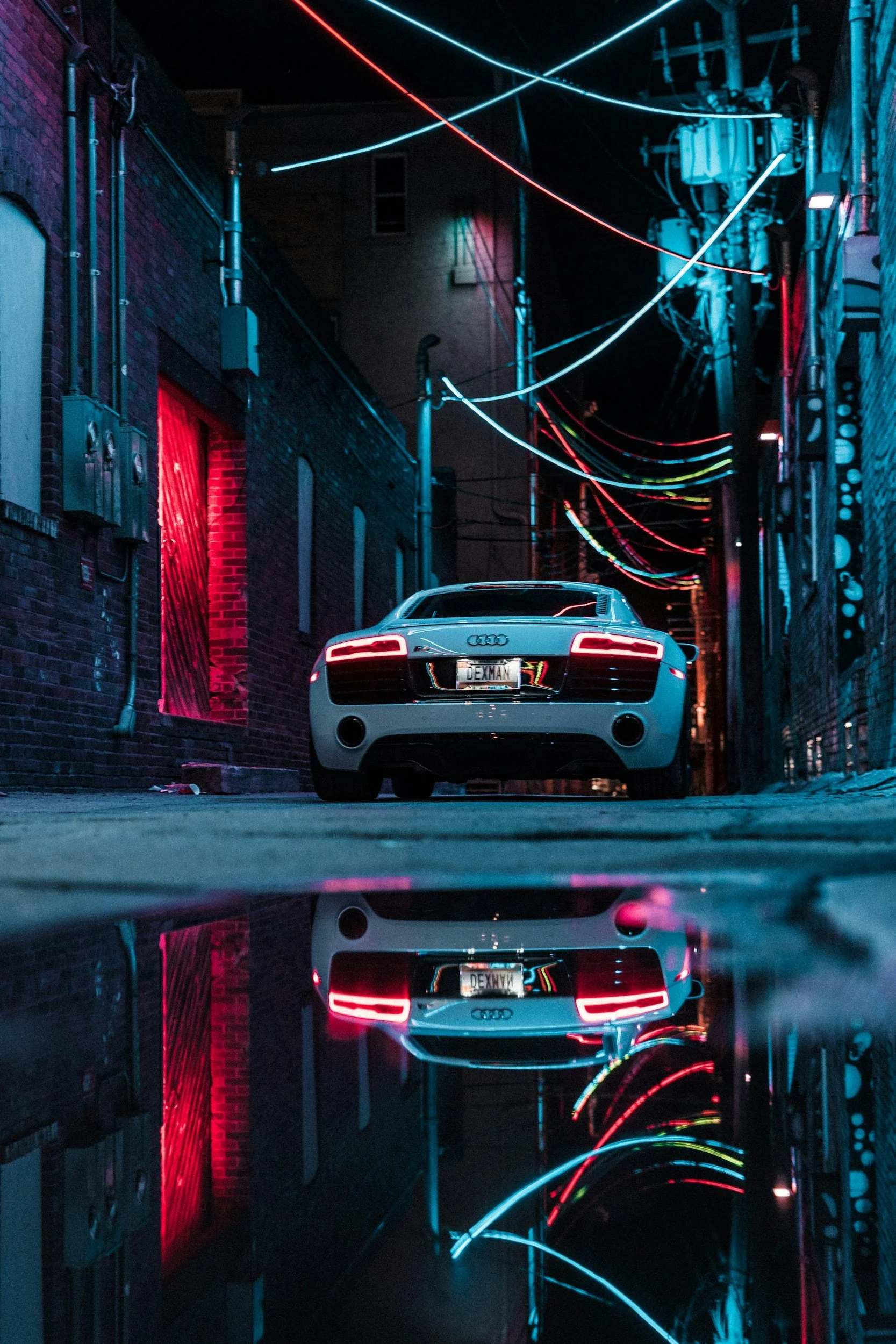 A white Audi sports car parked in a dark alley at night, illuminated by neon lights and reflections on wet pavement.