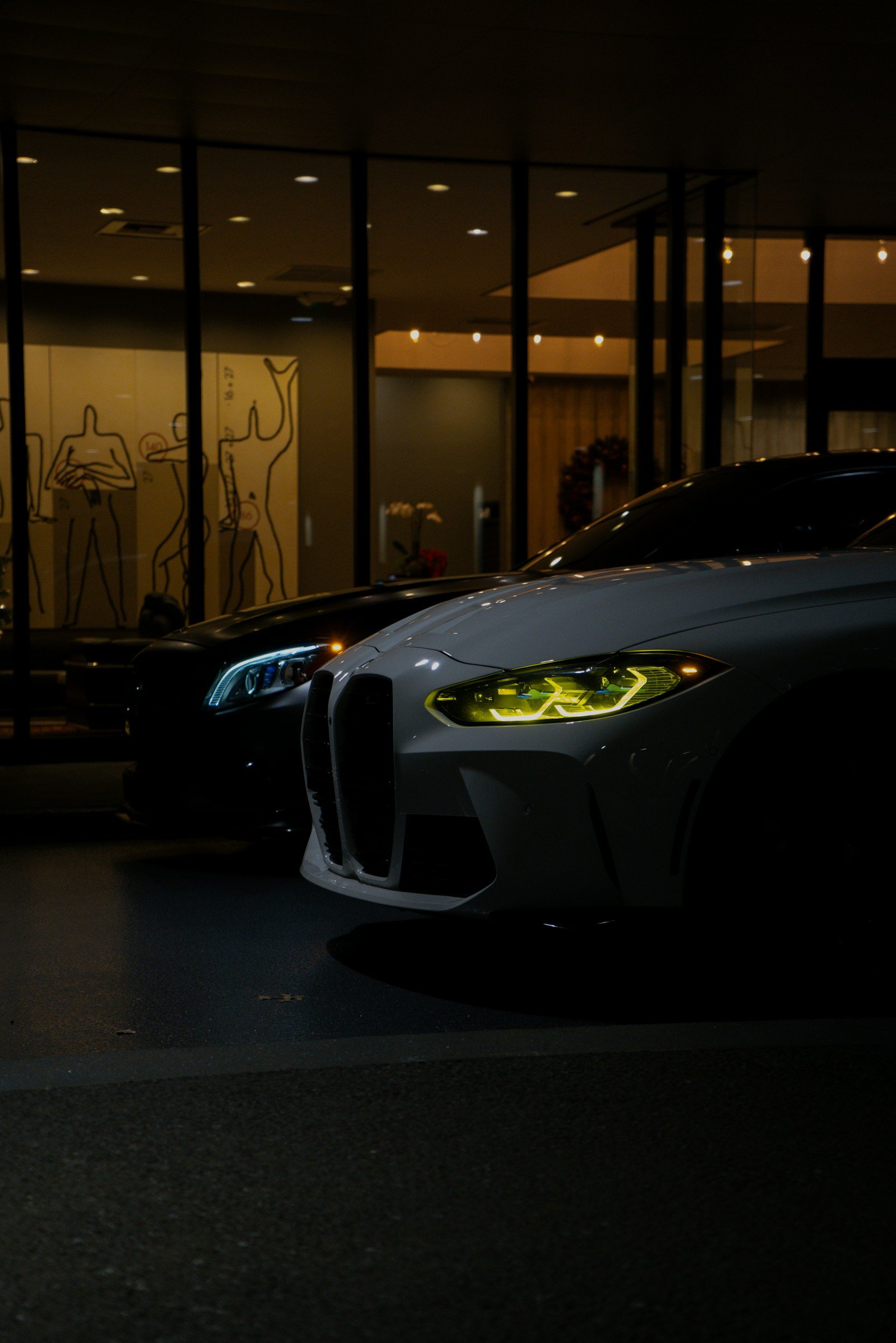 Nighttime shot of luxury cars parked outside a modern building with large glass windows and minimalistic wall art inside.