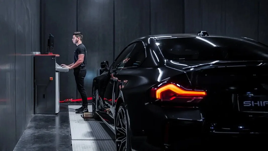 A man in black standing at a computer station inside a dark garage next to a black sports car with illuminated tail lights.
