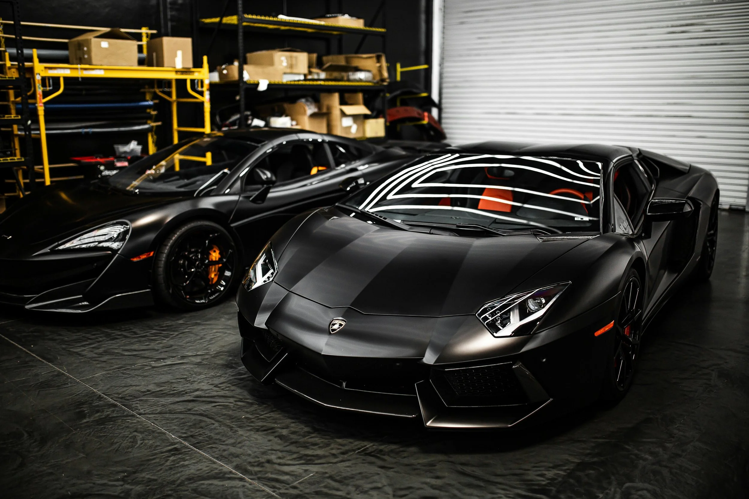 Two black luxury sports cars, a Lamborghini and a McLaren, parked inside a warehouse with black and yellow shelves and cardboard boxes in the background.