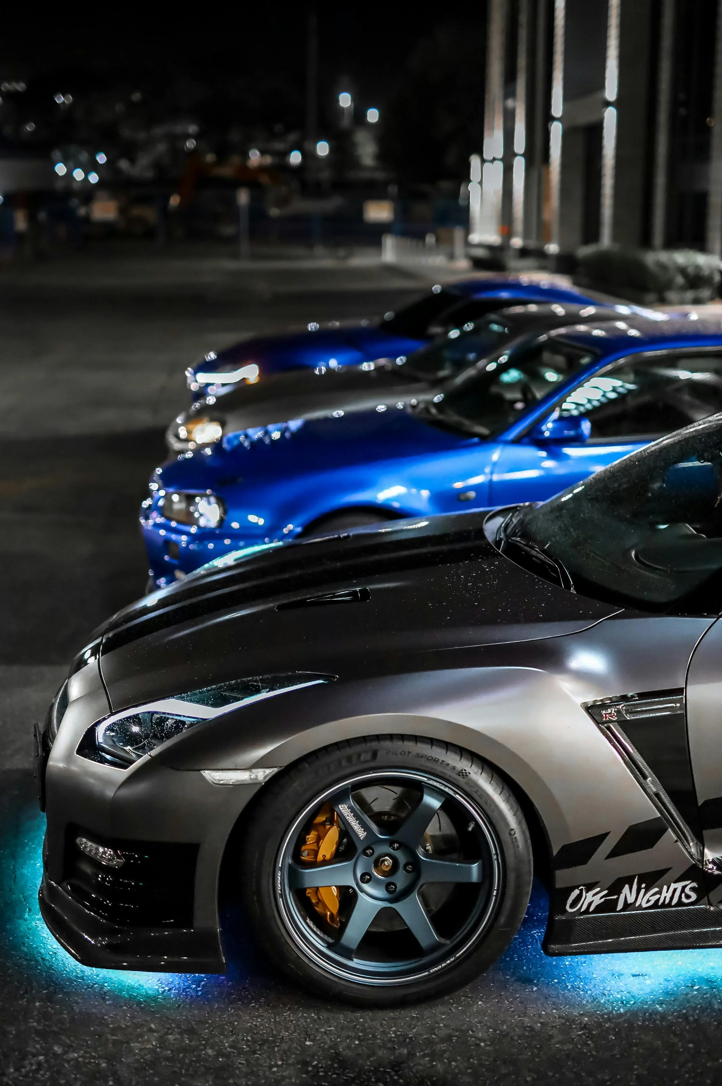 Close-up of several cars parked at night, including a black sports car with illuminated wheels and custom decals.