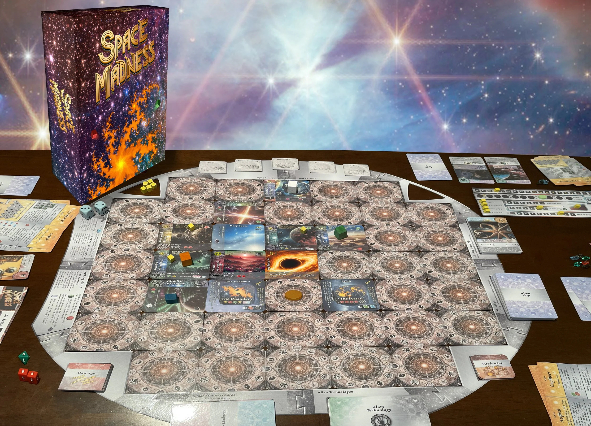 Photo of the board game Space Madness set up on a table with the game box, cards, tokens, and dice, against a starry space-themed backdrop.
