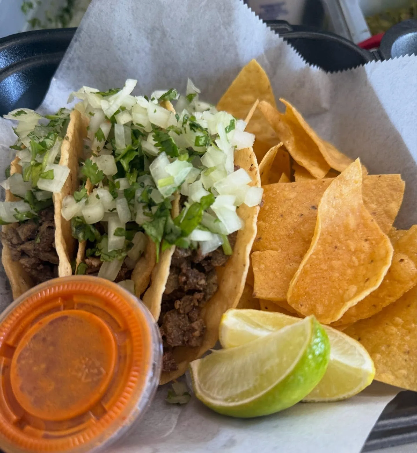Close-up of three beef tacos with chopped onions and cilantro, accompanied by tortilla chips, lime wedges, and a small container of salsa or hot sauce.