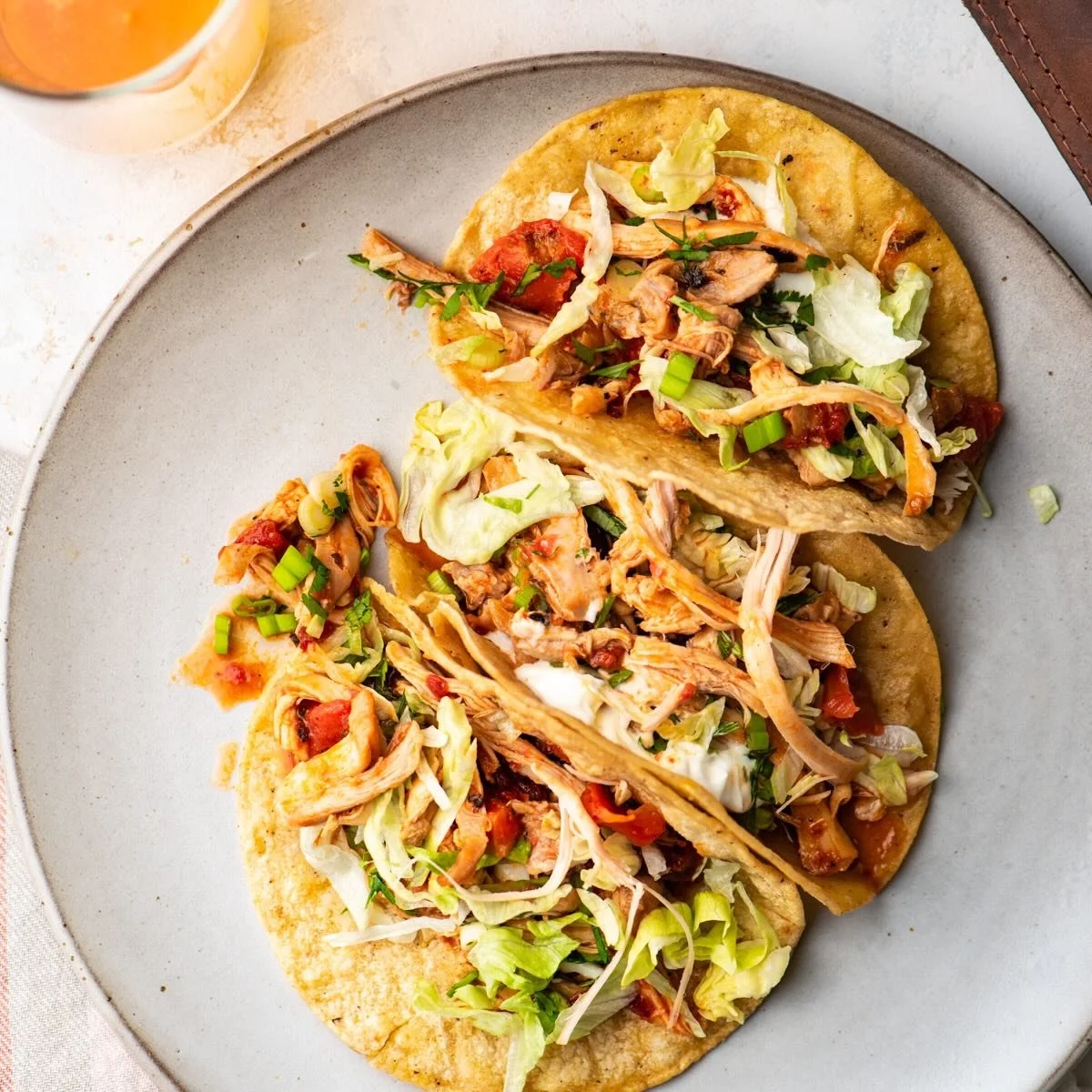 Three shredded chicken tacos with lettuce, tomatoes, and green onions on a round gray plate.