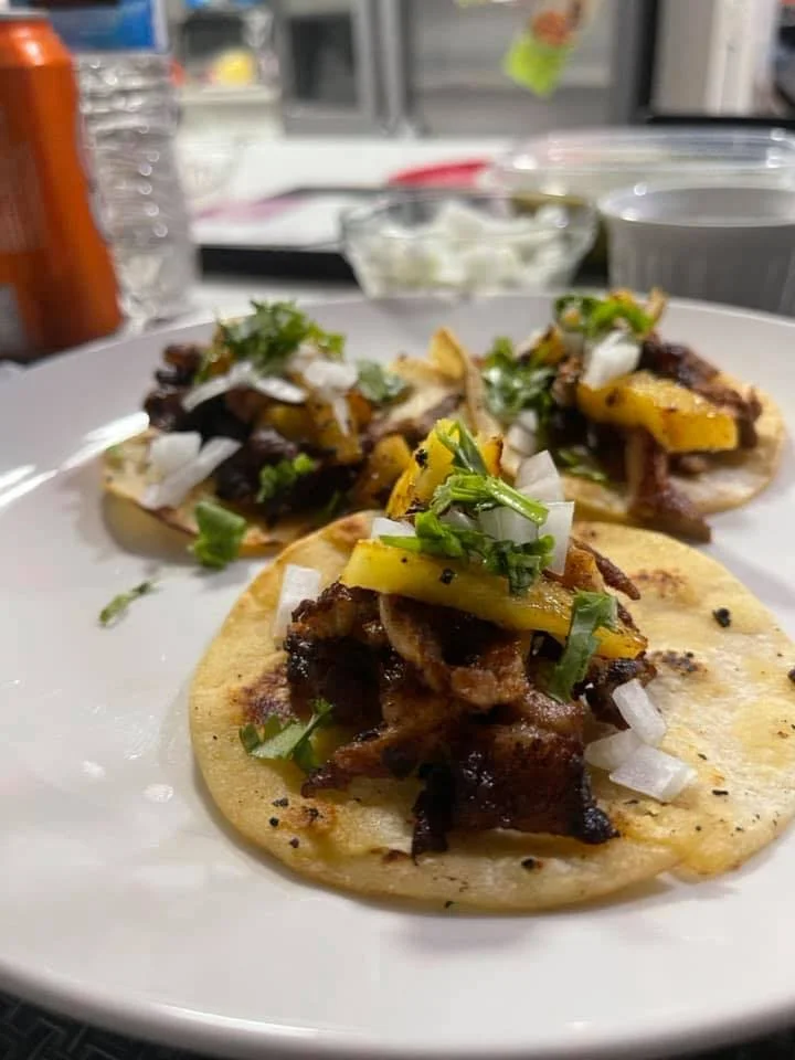Close-up of tacos with grilled meat, topped with chopped onions, cilantro, and sliced yellow peppers on a white plate.