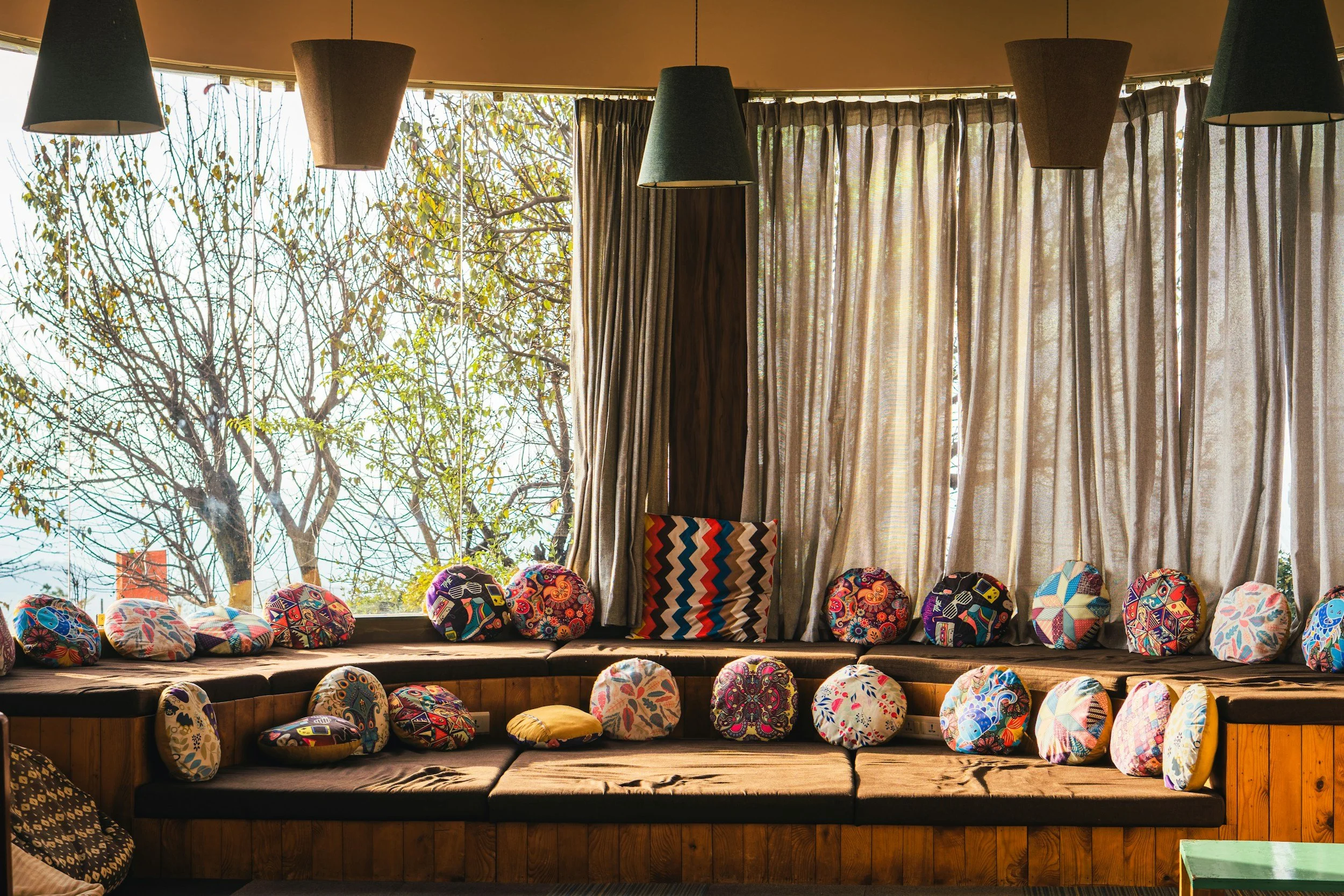 A cozy seating area with a large window, lined with colorful, patterned cushions and pillows, some on the couch and some on the floor. The window has beige curtains, and outside there are leafless trees and a clear blue sky.