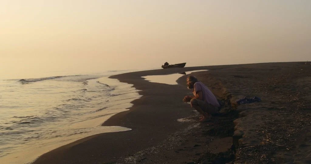 A man sitting on the sandy beach near the water, watching the sunset with a boat anchored in the distance.