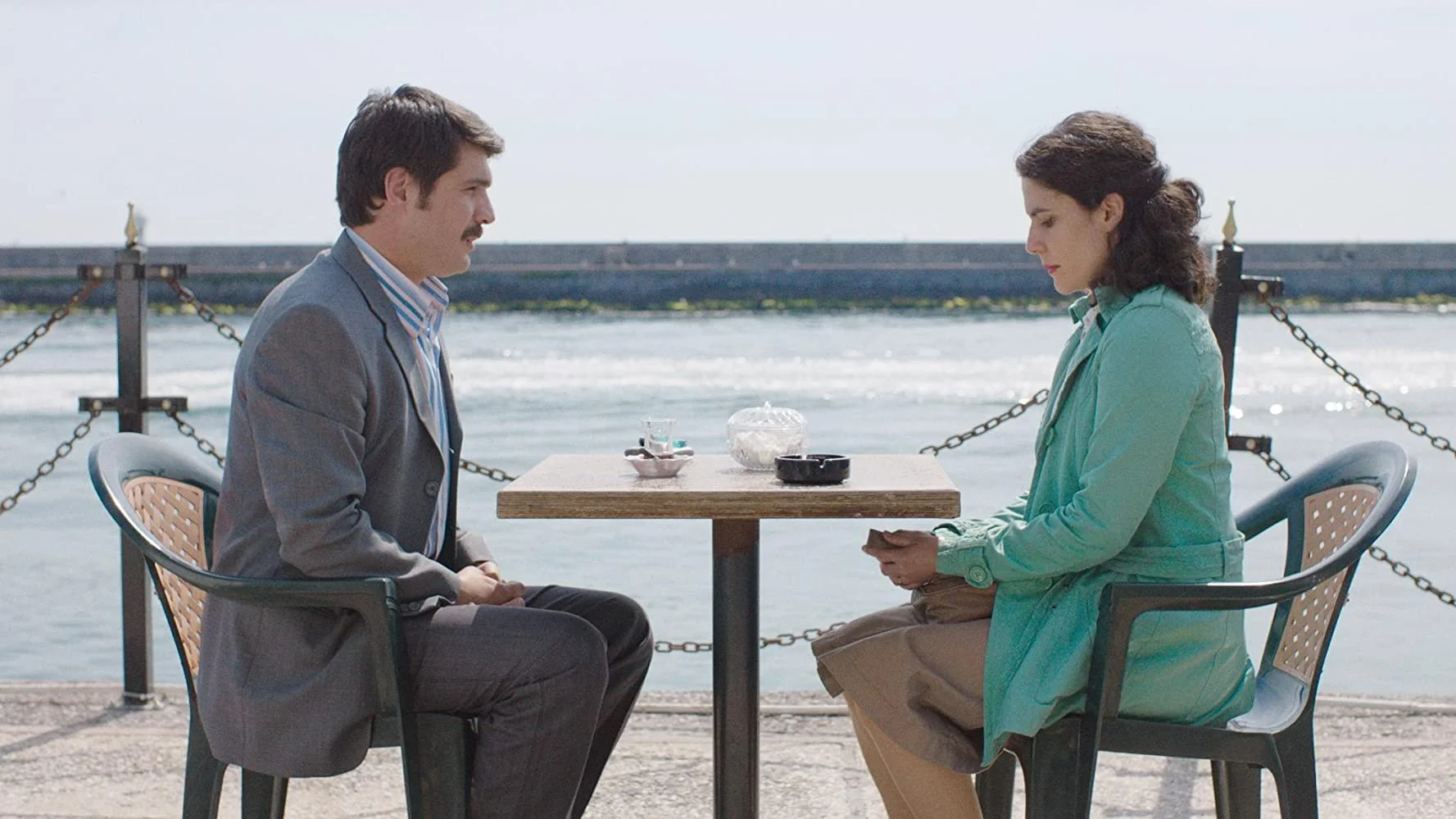 A man and woman sitting across from each other at a table near the water, with a chain-link fence and water in the background.