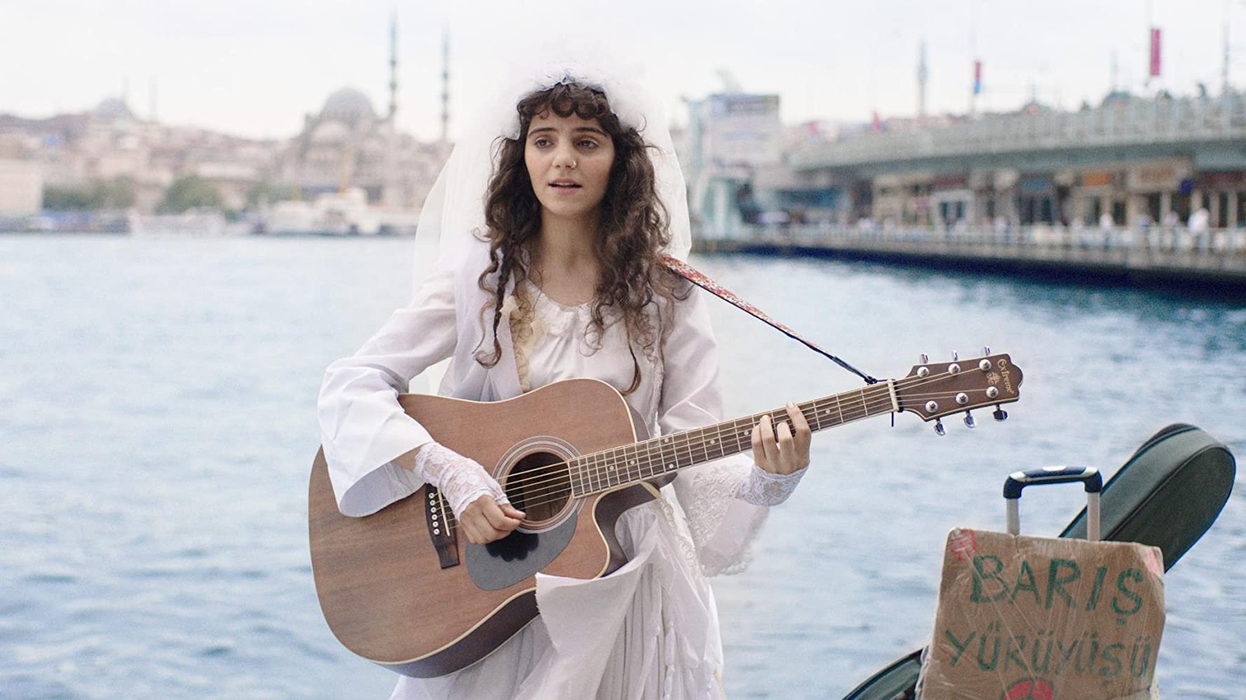 A young woman dressed in white with a veil, playing an acoustic guitar by a waterfront, with a cityscape and bridge in the background, and a cardboard sign with writing in Turkish near her foot.