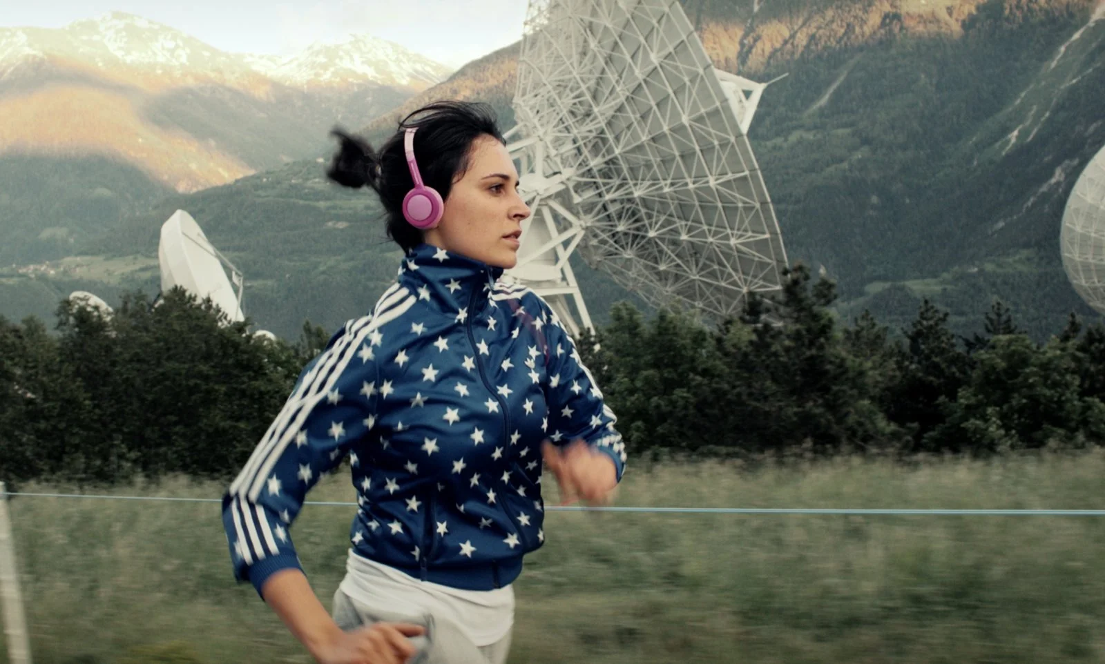 A woman jogging outdoors with a mountain and multiple satellite dishes in the background. She is wearing pink headphones and a blue jacket with white stars.
