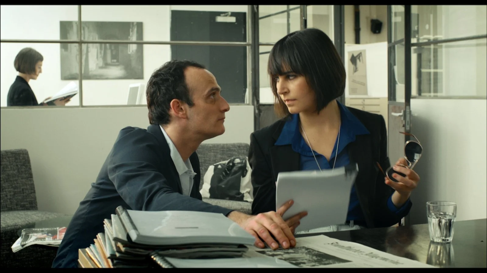 A man and woman sitting at a table in an office, engaging in an intense conversation, with documents and folders on the table and one holding a tablet and sunglasses, while a woman reads a book in the background.