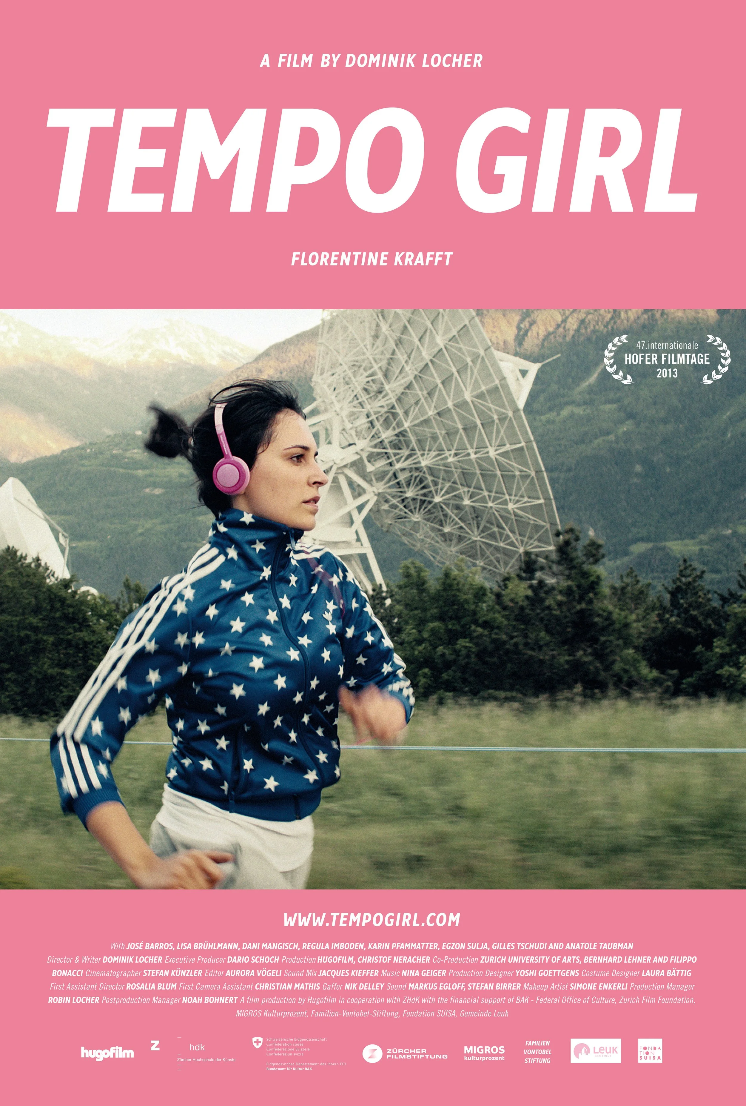 A woman jogging outdoors wearing a blue jacket with white stars and pink headphones. There is a large satellite dish and mountainous landscape in the background. Promotional poster for the film "Tempo Girl" by Dominik Locher.