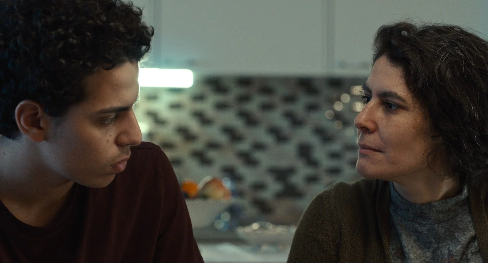 Two women having a serious conversation indoors, with a blurred patterned background.