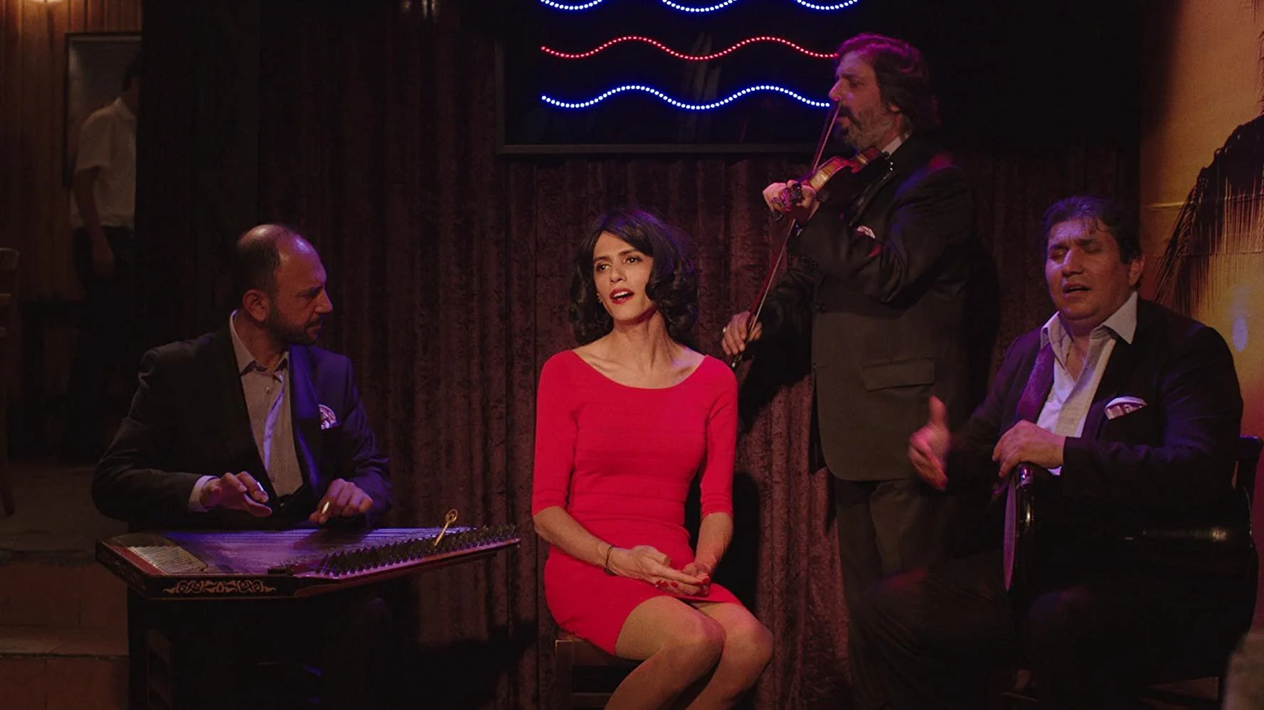 A woman in a red dress is performing on stage with three men, one playing a musical instrument, another playing a violin, and a third sitting and possibly singing, in a dimly lit venue with brown curtains and neon lights above.