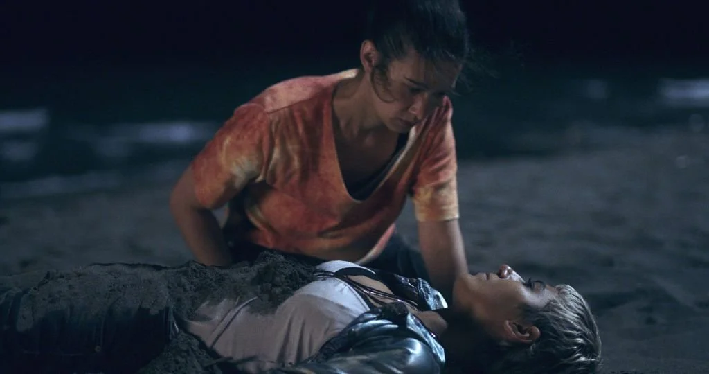 Two women on a dark beach at night, one woman is lying on the sand while the other kneels beside her, holding her and looking at her with concern.