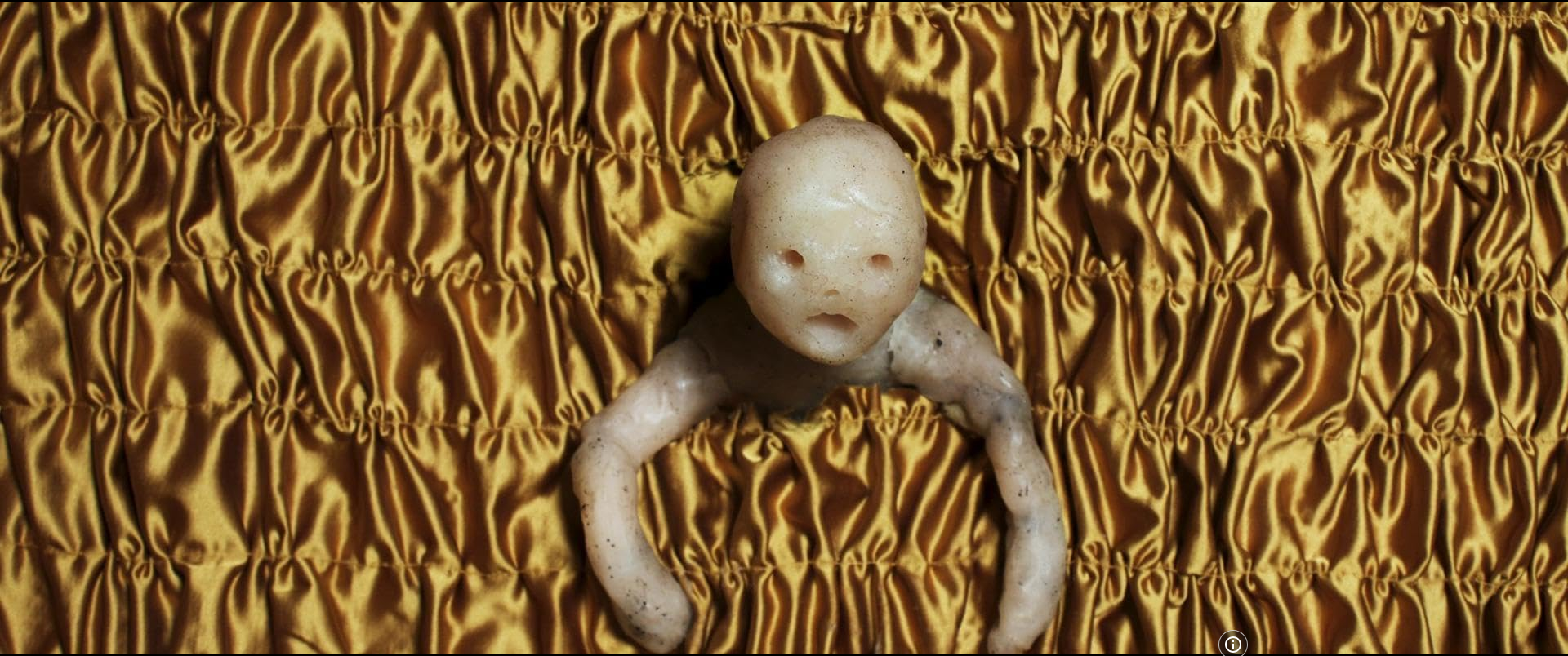 A baby doll with a human face and body, lying on a gold-colored, ruched fabric background.