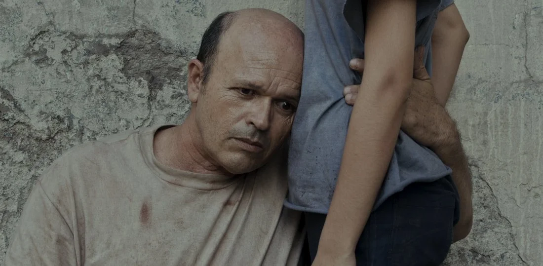 A man with a distressed expression leaning against a weathered wall, with a child sitting on his lap, partly visible.