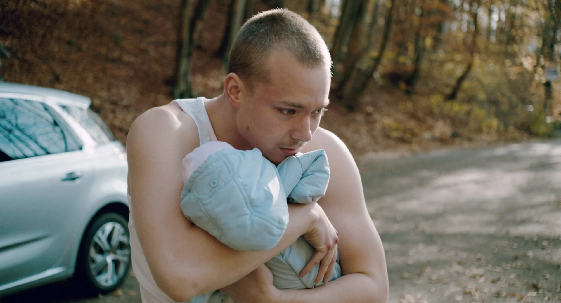 A shirtless young man with short hair hugging a small child wrapped in a light blue blanket outdoors near a silver car and trees with autumn foliage.